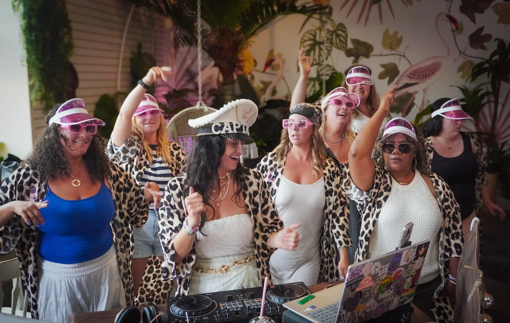 Group of women dancing and celebrating at a party, wearing pink visors, leopard-print jackets, and white dresses, with a DJ setup and laptops in the foreground.