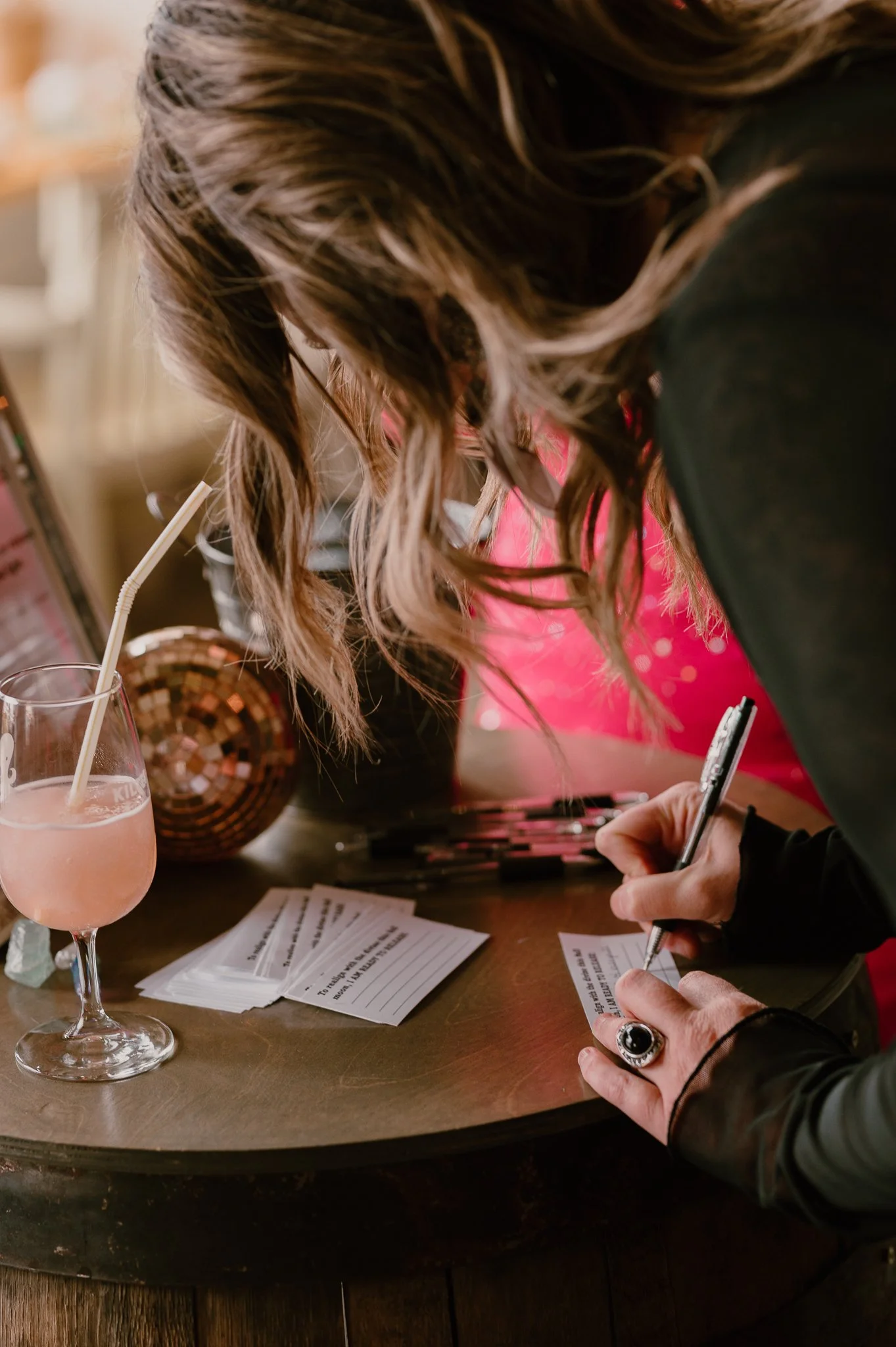 A woman with wavy brown hair signing a card on a wooden table with a pink drink, makeup products, and handwritten notes.