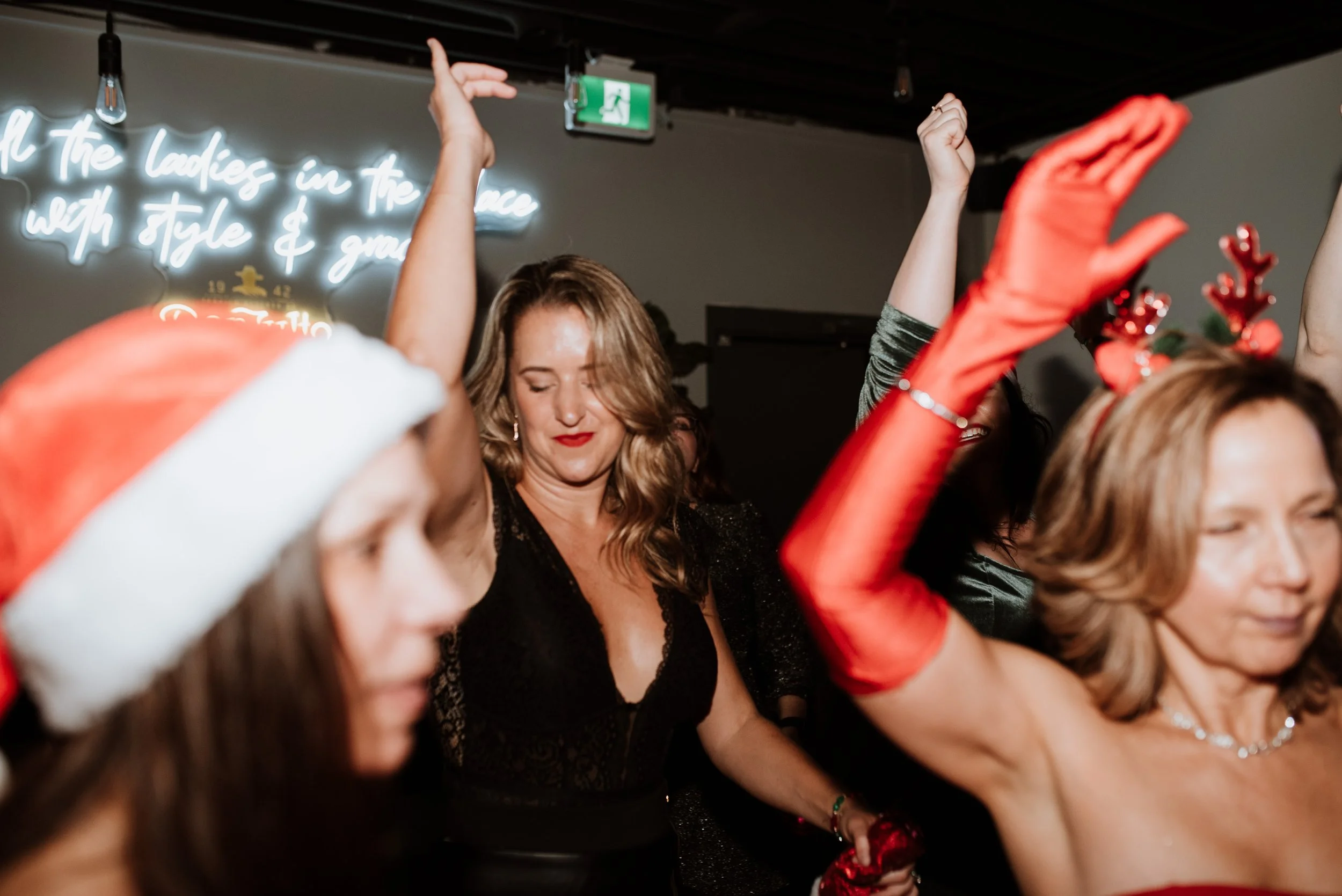 Women dancing and celebrating at a Christmas party, some wearing holiday accessories like a Santa hat and reindeer antlers, with a neon sign in the background.