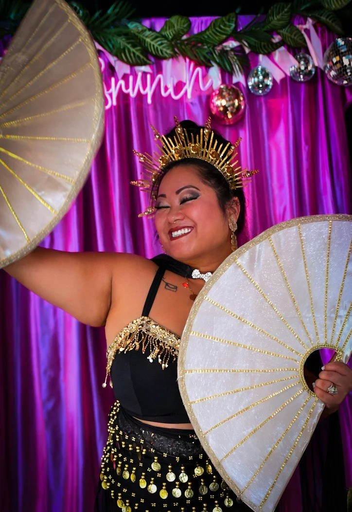A woman dressed in a black and gold costume, wearing a golden crown, holding two fans, with a vibrant purple curtain and decorative greenery in the background, smiling joyfully.