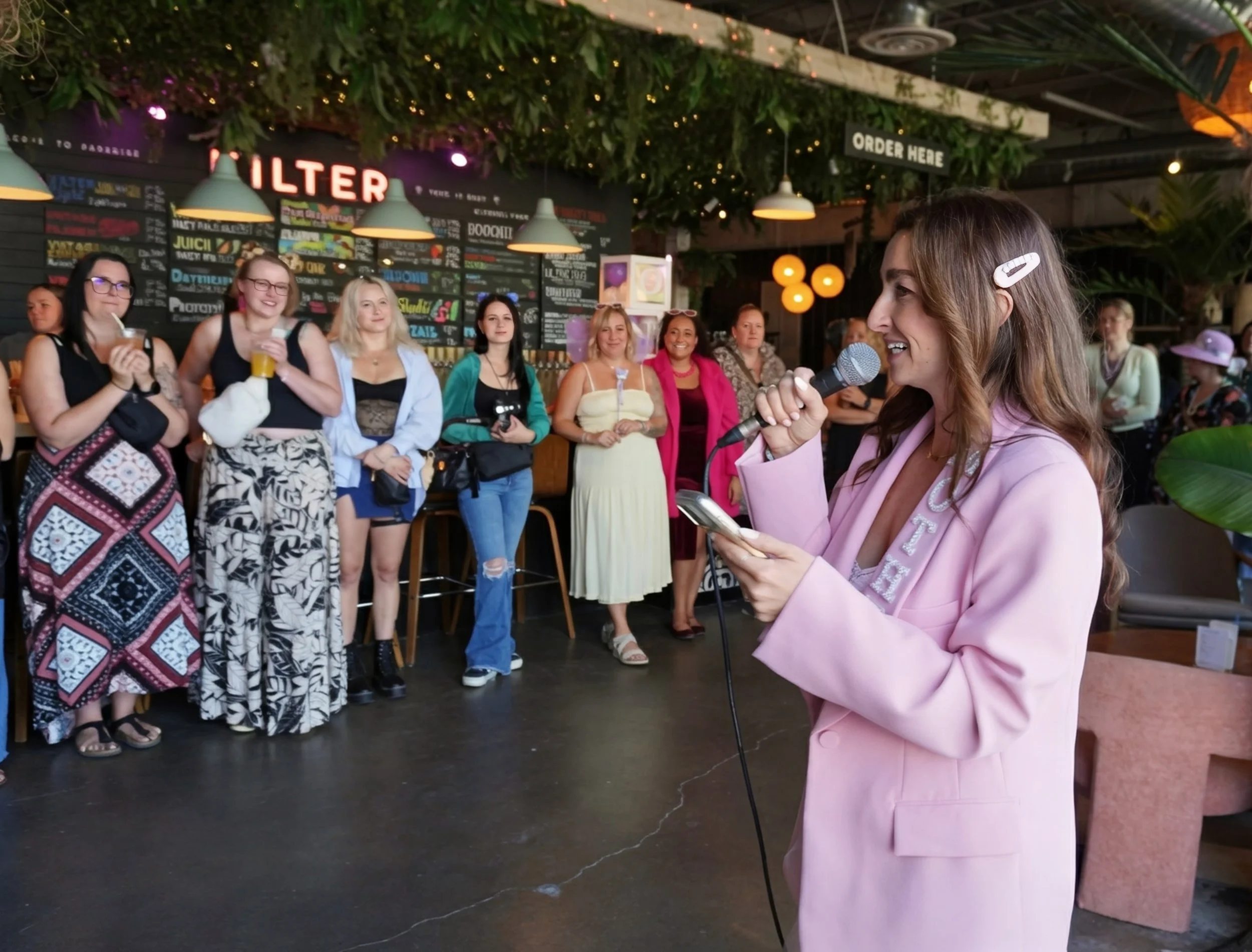 A woman in a pink blazer with a hair clip is speaking into a microphone and reading from her phone in front of a group of diverse women in a restaurant or bar with a menu on the wall behind them.