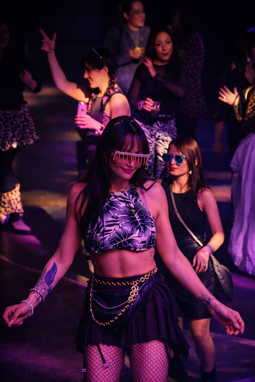 A group of women dancing at a party. The woman in the foreground is wearing a tropical print crop top, fishnet stockings, and fashionable jewelry with a chain belt and glasses. Other women in the background are in dark and patterned clothing, holding drinks and enjoying the event.