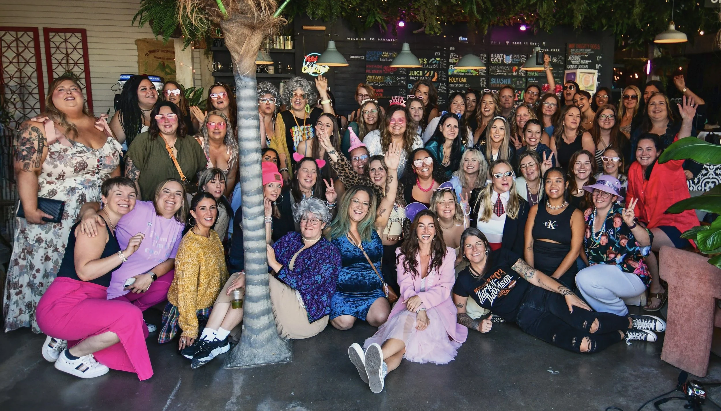 A large group of women gathered indoors for a celebration, smiling and posing for a group photo, with some making peace signs and wearing festive accessories.