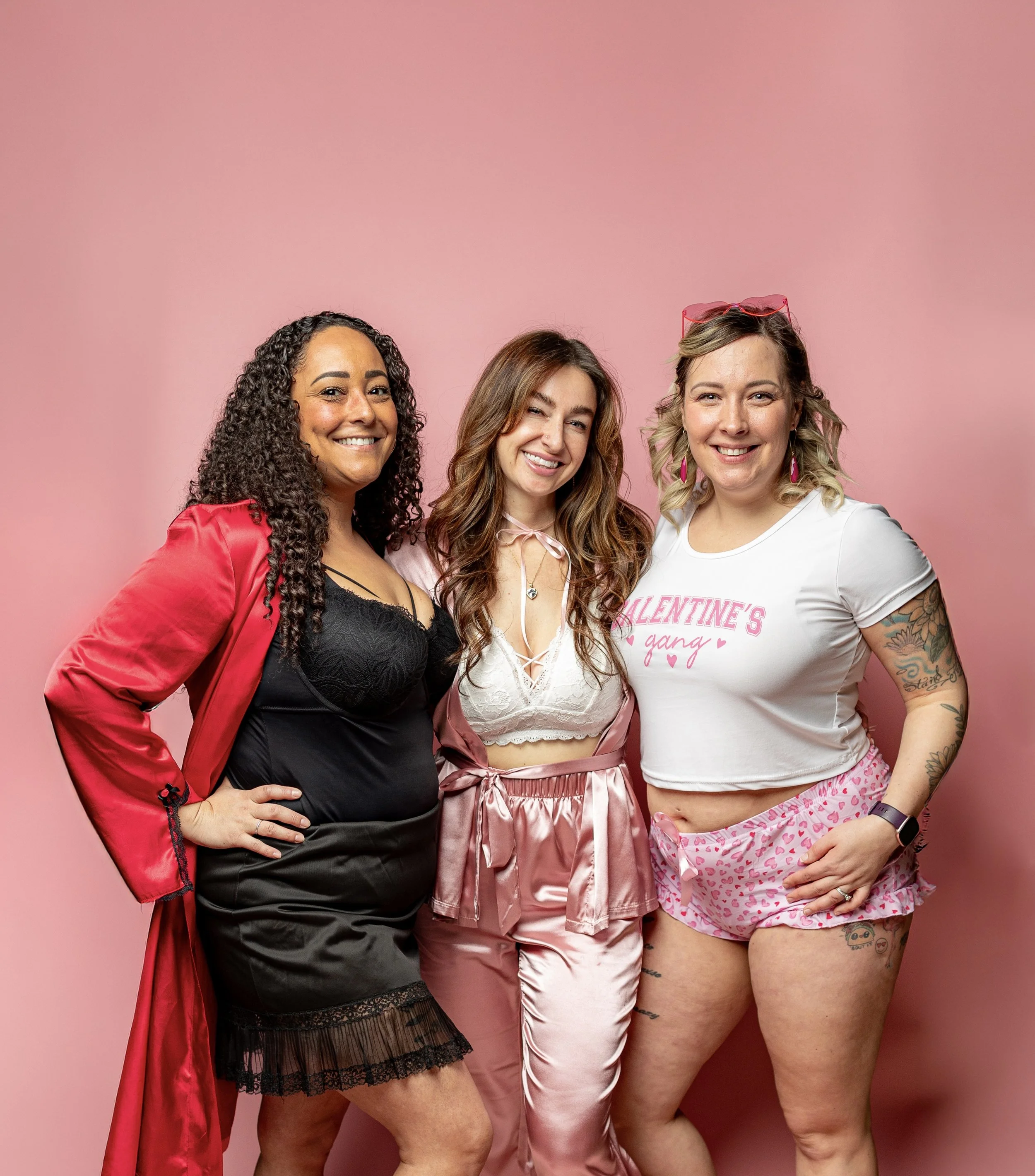 Three women standing together against a pink background, dressed in lingerie and casual clothing, smiling at the camera.