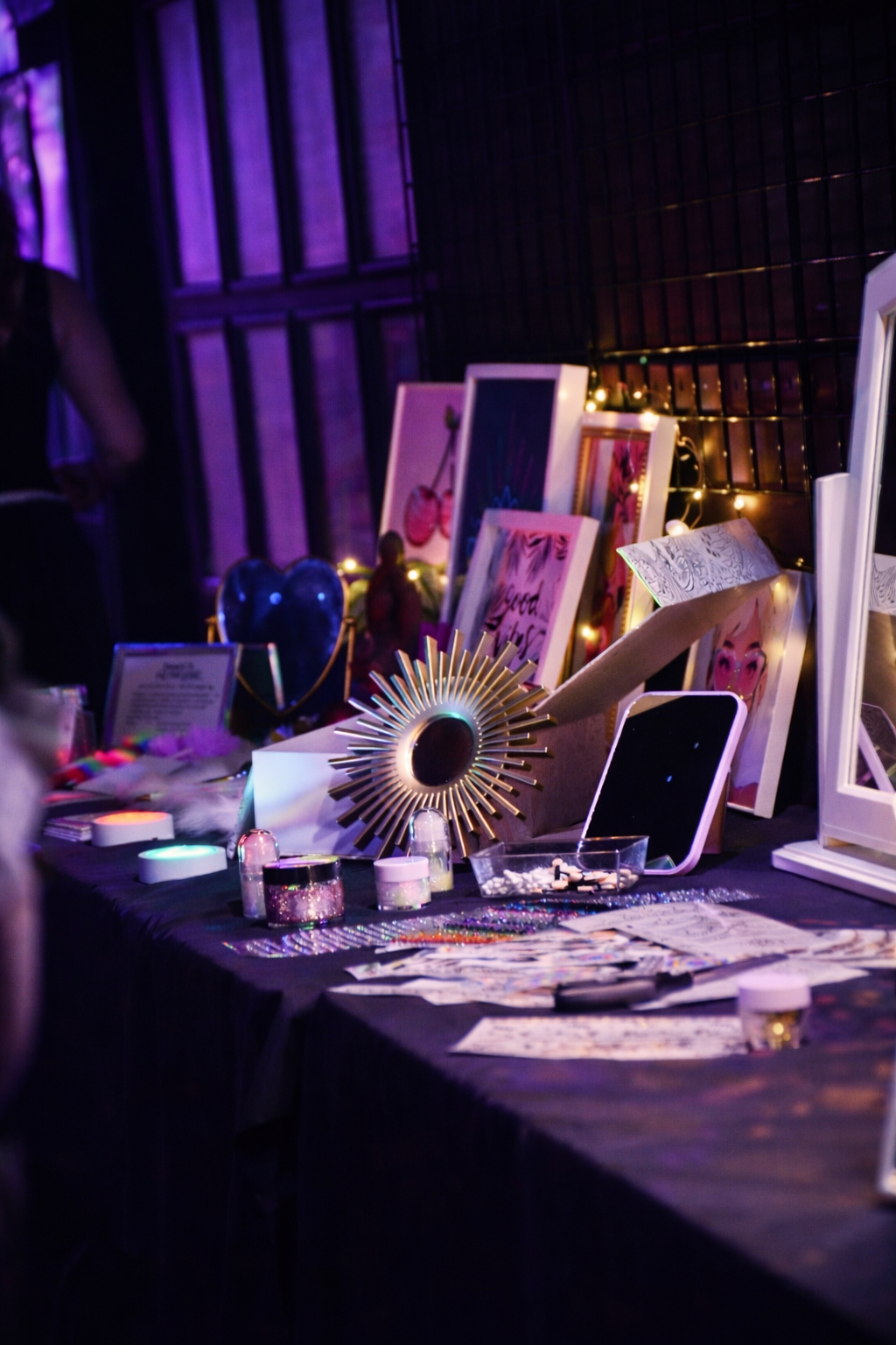 A display table with various framed artwork, decorative items, and colorful jewelry against a dark background illuminated by purple and fairy lights.