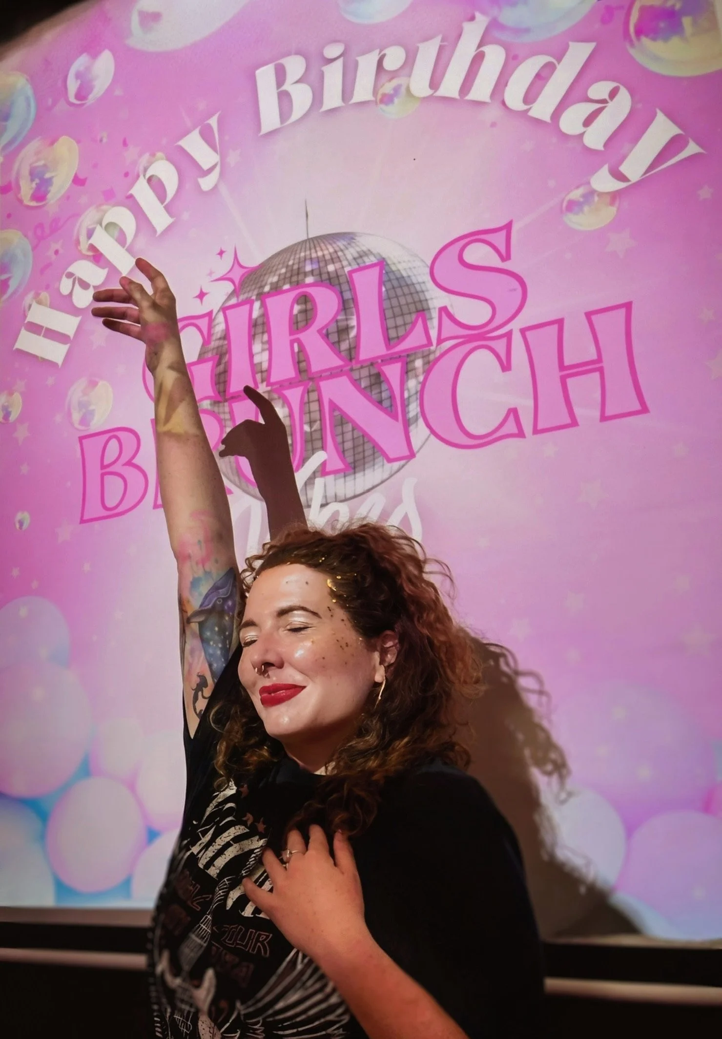 A woman with curly hair, red lipstick, and glitter on her face, smiling and posing with one arm raised in front of a pink birthday background decorated with a disco ball, balloons, and the words 'Happy Birthday Girls Launch'.