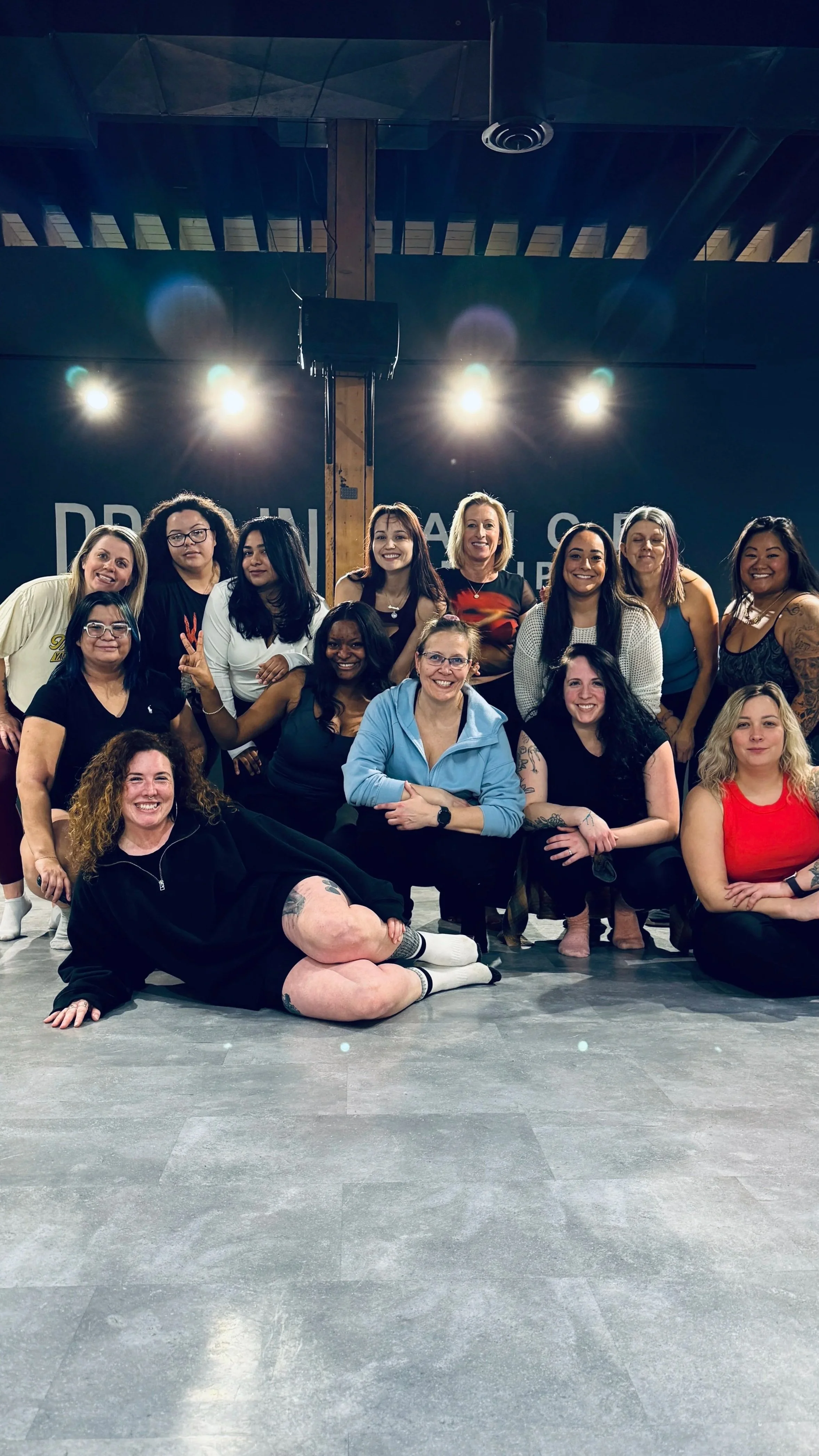 A group of women posing together in a studio with stage lights overhead and a black background that has partially visible white text.