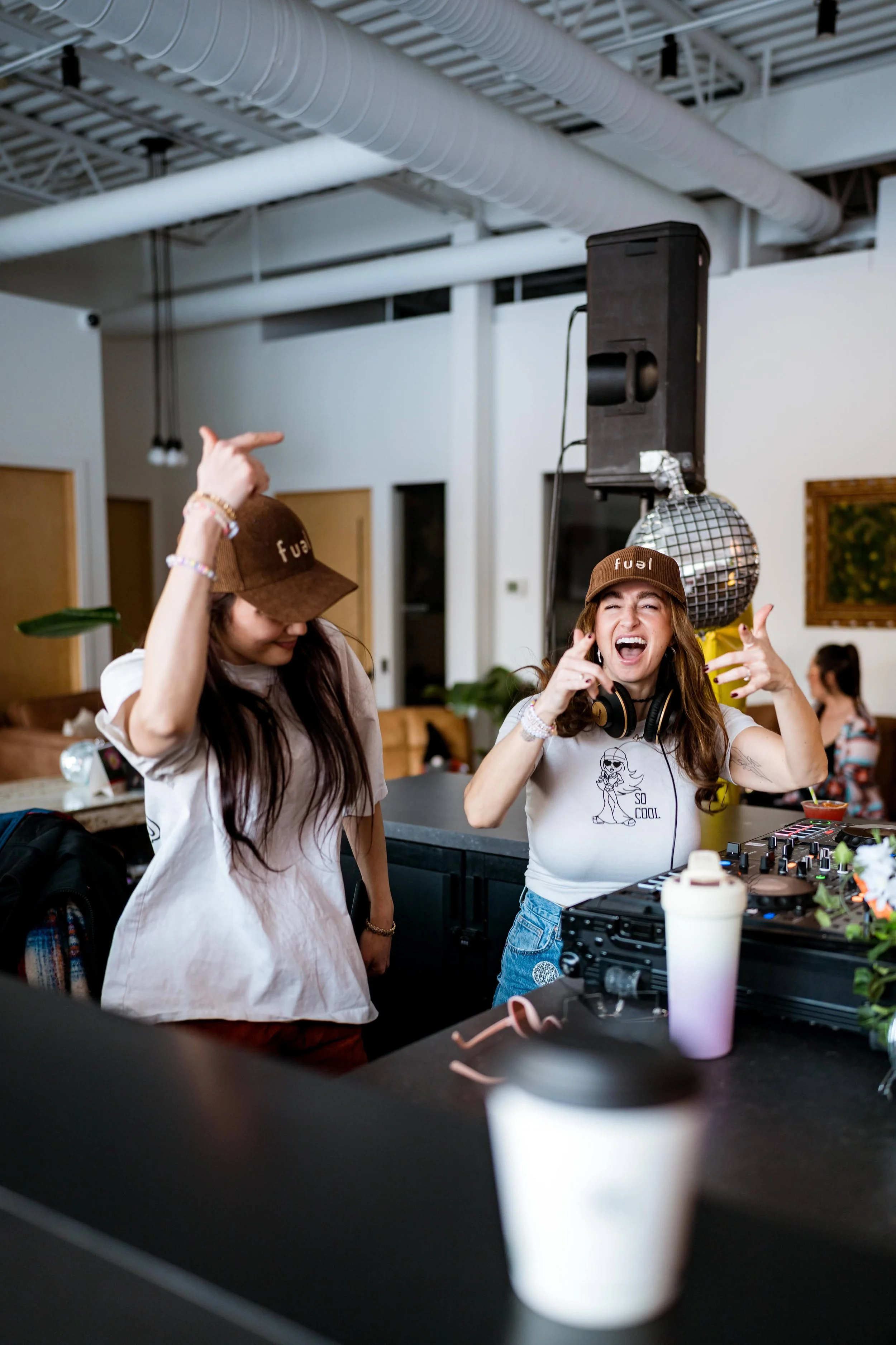 Two women wearing brown hats and casual clothing dancing and singing at a DJ booth in a lively indoor setting.