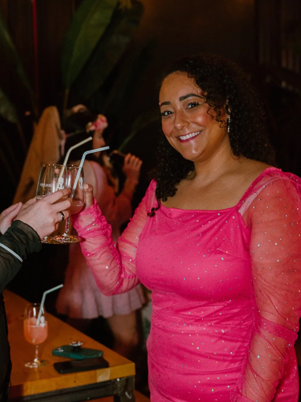 Woman in a pink dress smiling and holding a drink with a straw at a social gathering.