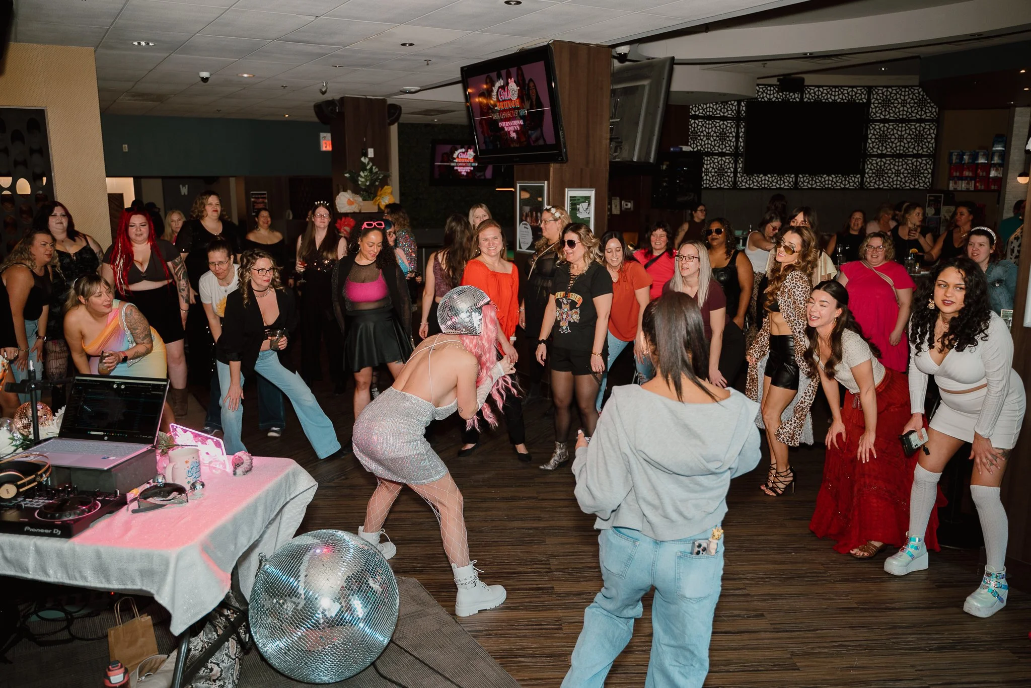 A woman with pink hair and a silver dress performs in front of a group of people at a dance party, with a disco ball on the floor and a DJ setup on a table nearby.