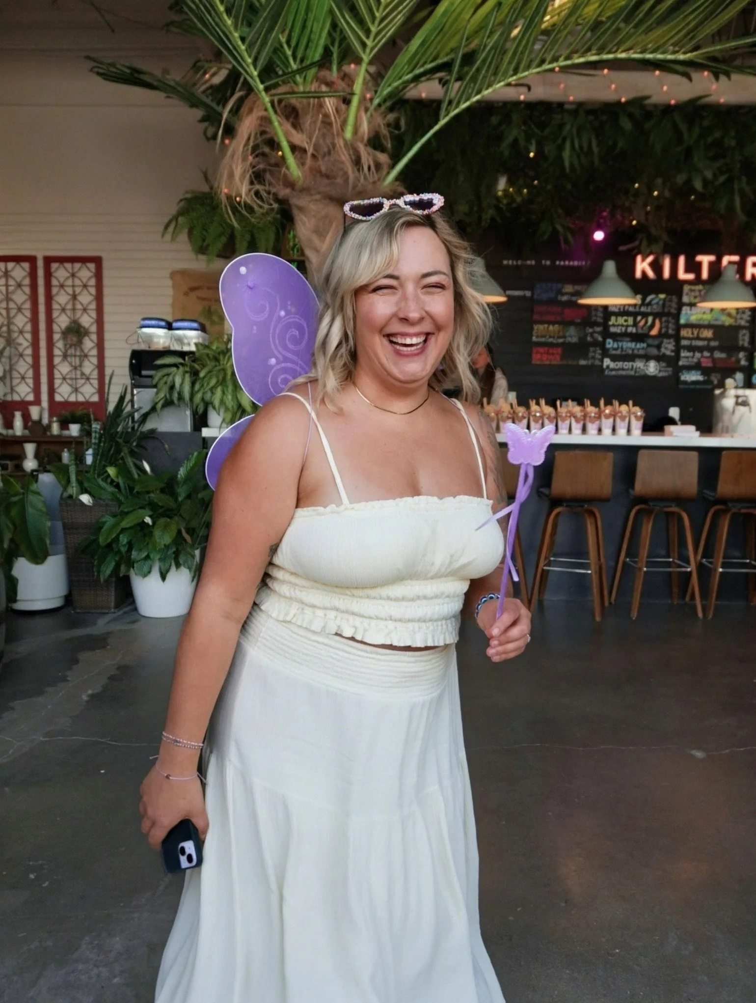 A woman dressed in a white dress with fairy wings and a flower wand, smiling and holding a phone, standing inside a cafe with plants and a blackboard menu in the background.