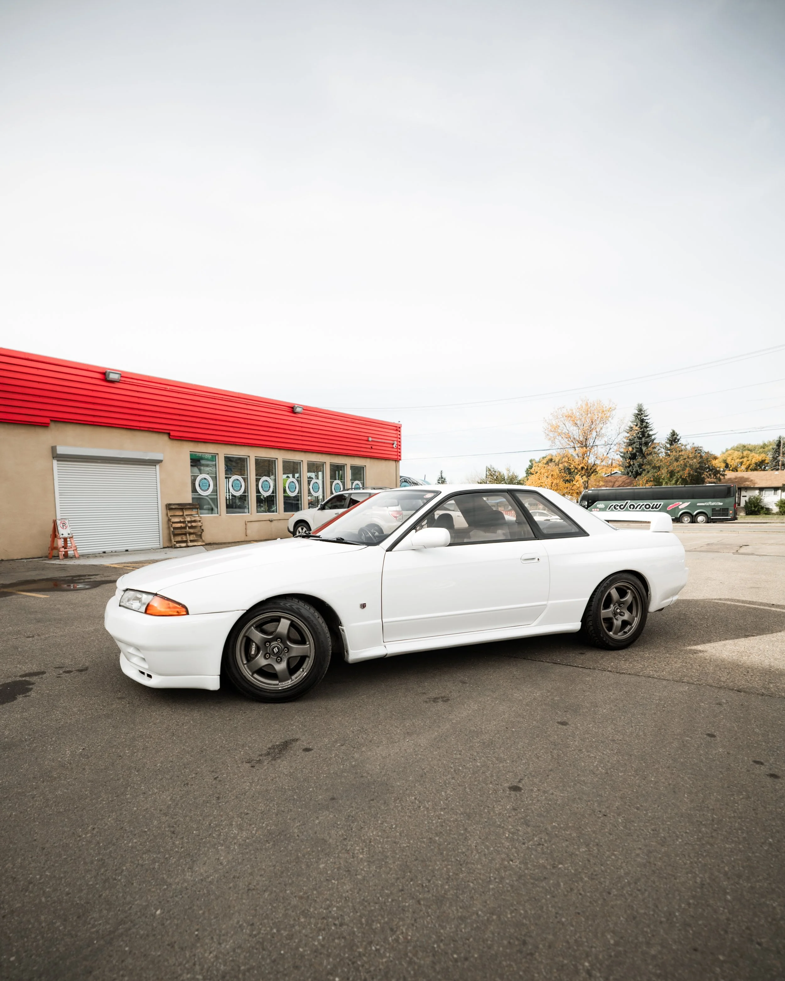 A white coupe car parked in a parking lot near a building with a red roof and large windows, with a bus and trees in the background.