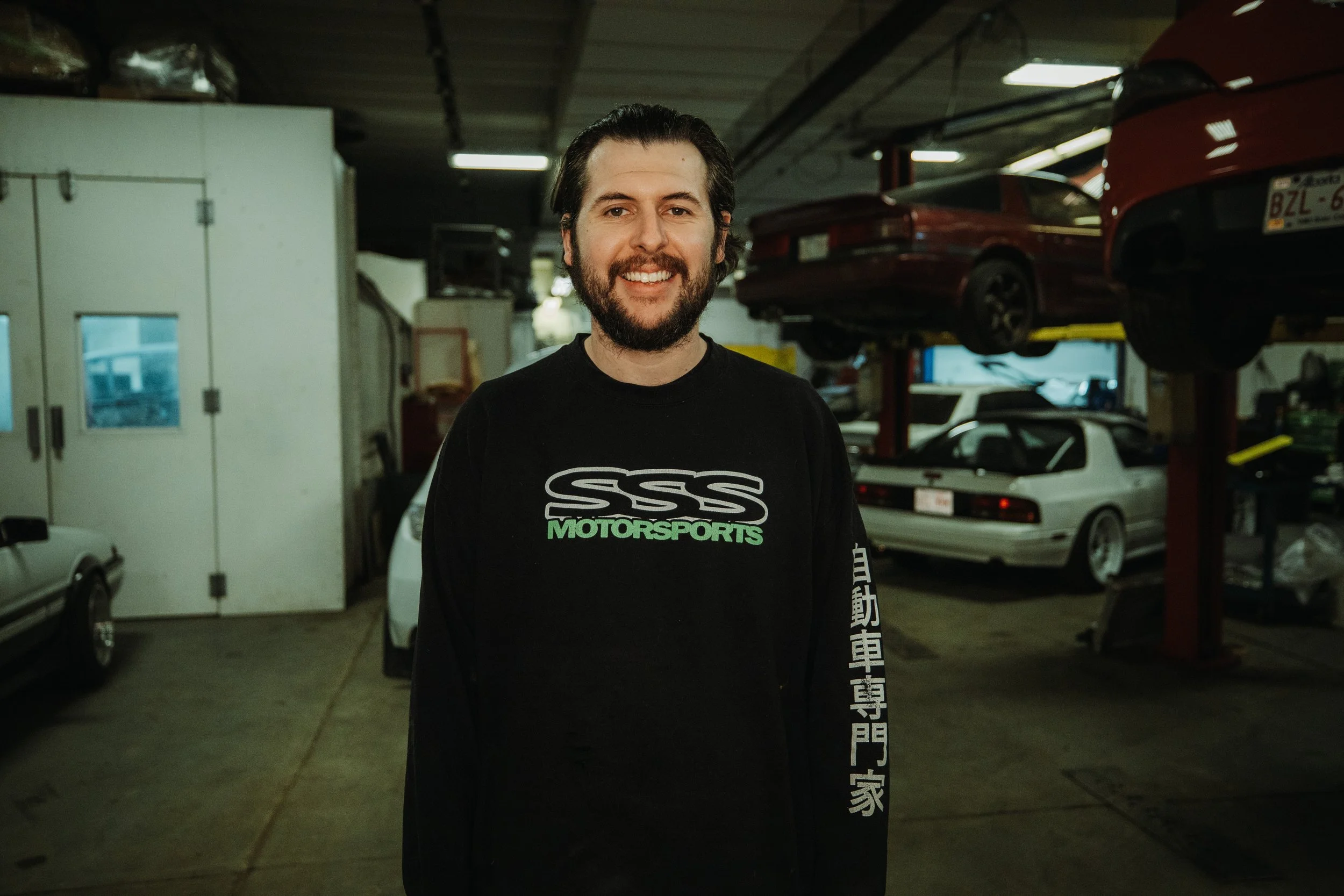 A man with dark hair and a beard smiling, standing in a garage with cars on lifts in the background.