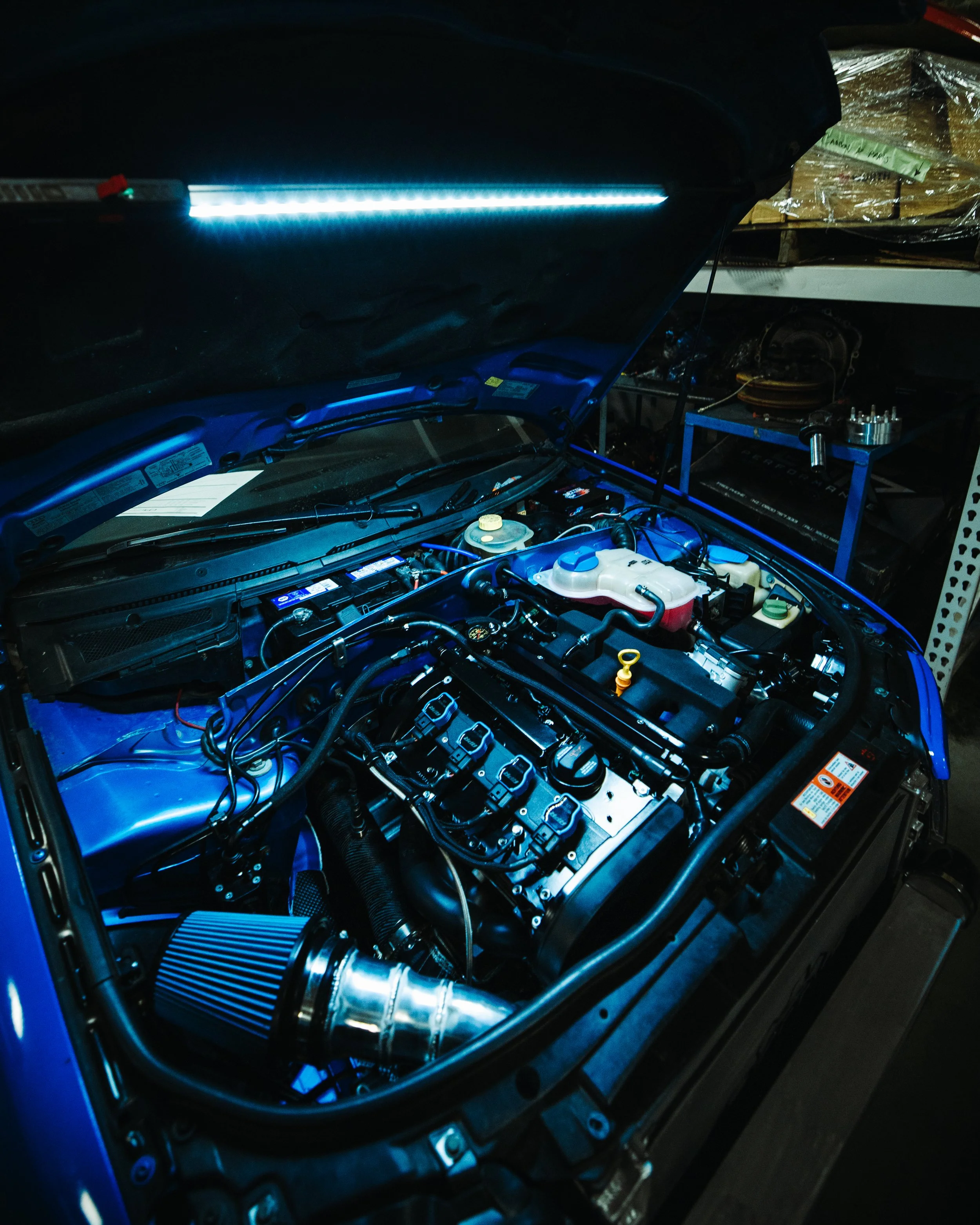 Under the hood of a blue sports car with LED lighting, showing a high-performance engine with various components and a cone-shaped air filter.