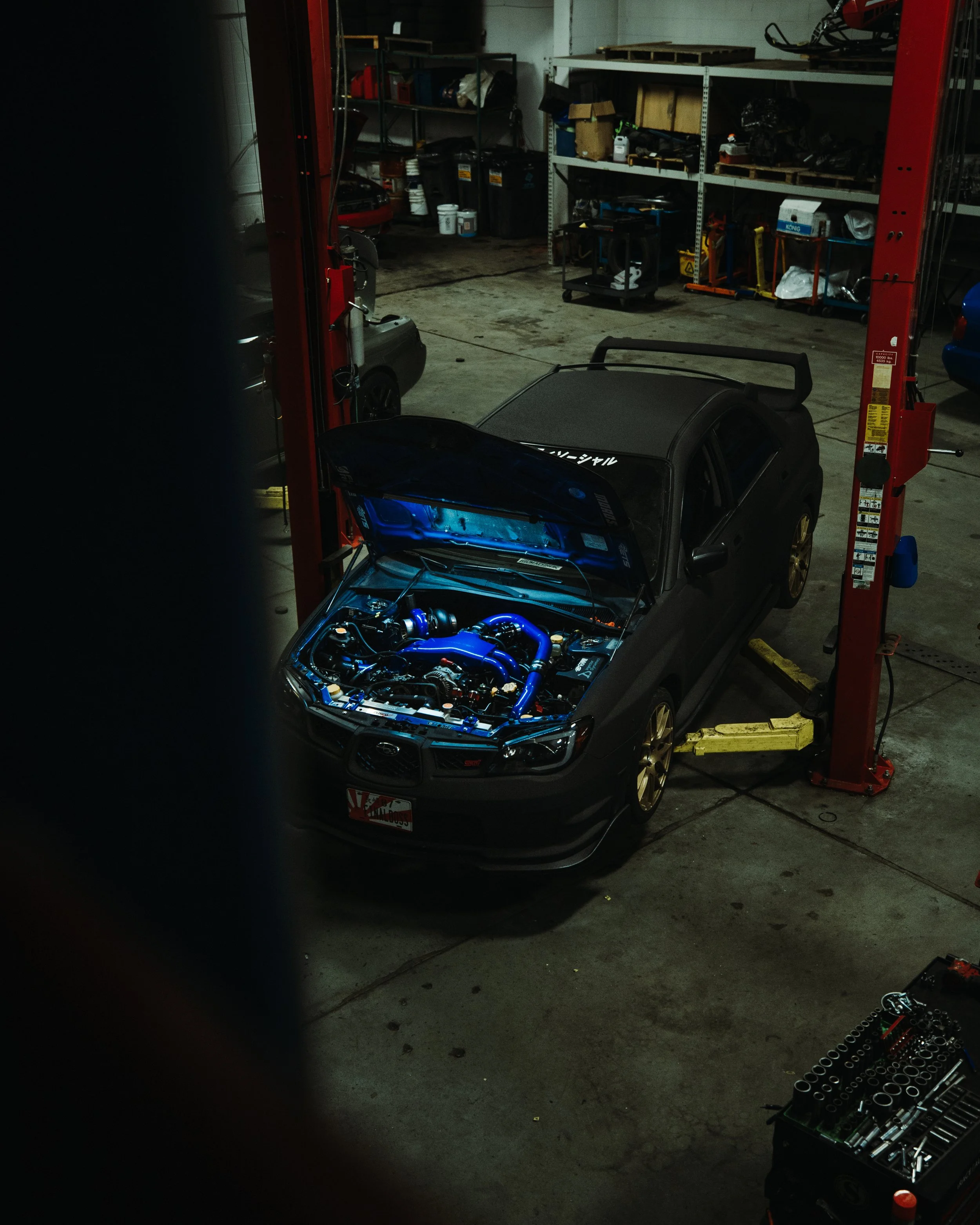 A dark-colored car with its hood open showing a modified engine with blue hoses, in a garage or workshop setting with shelves and tools in the background.