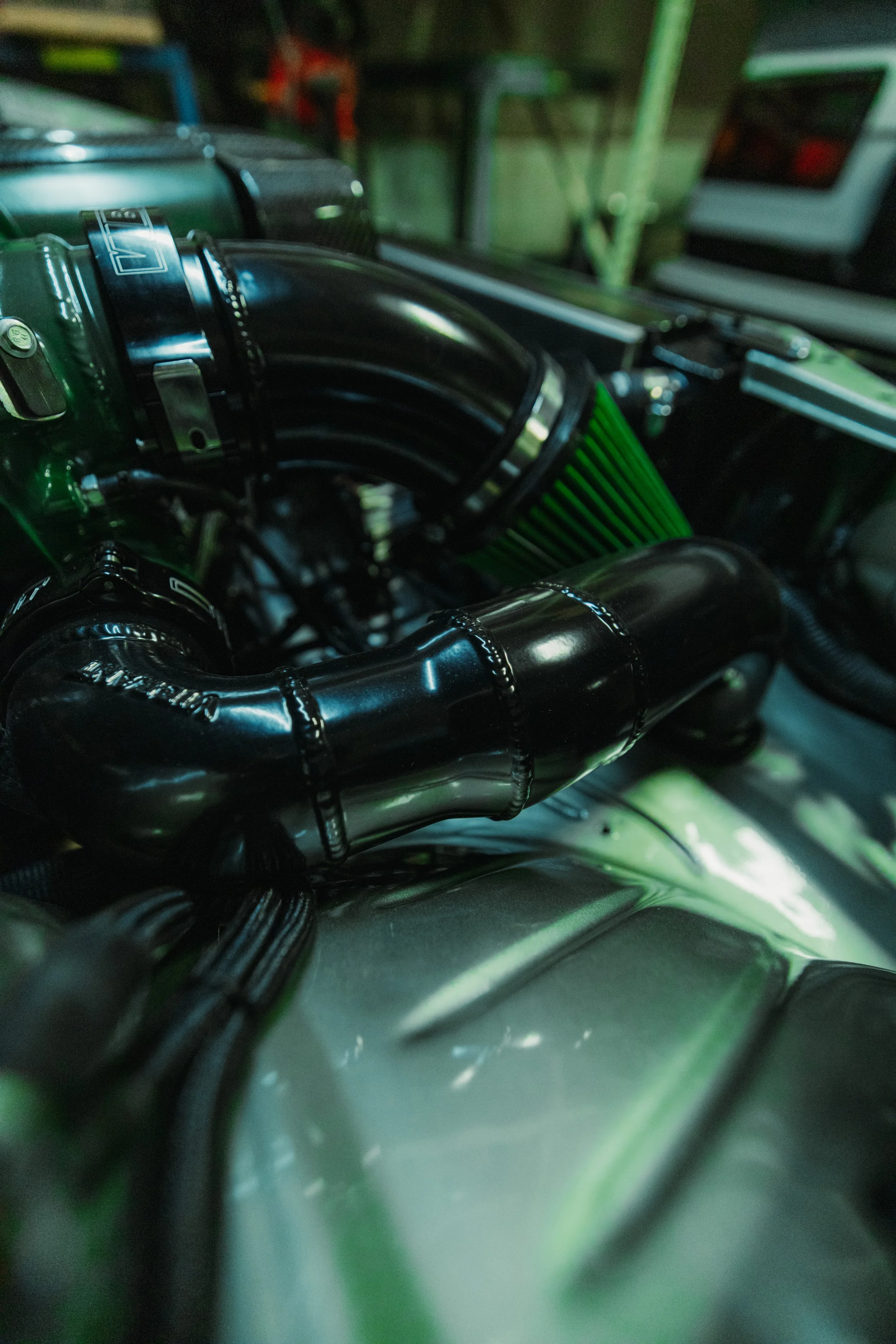 Close-up of a car engine with black pipes and green air filter, in a dimly lit garage or workshop.