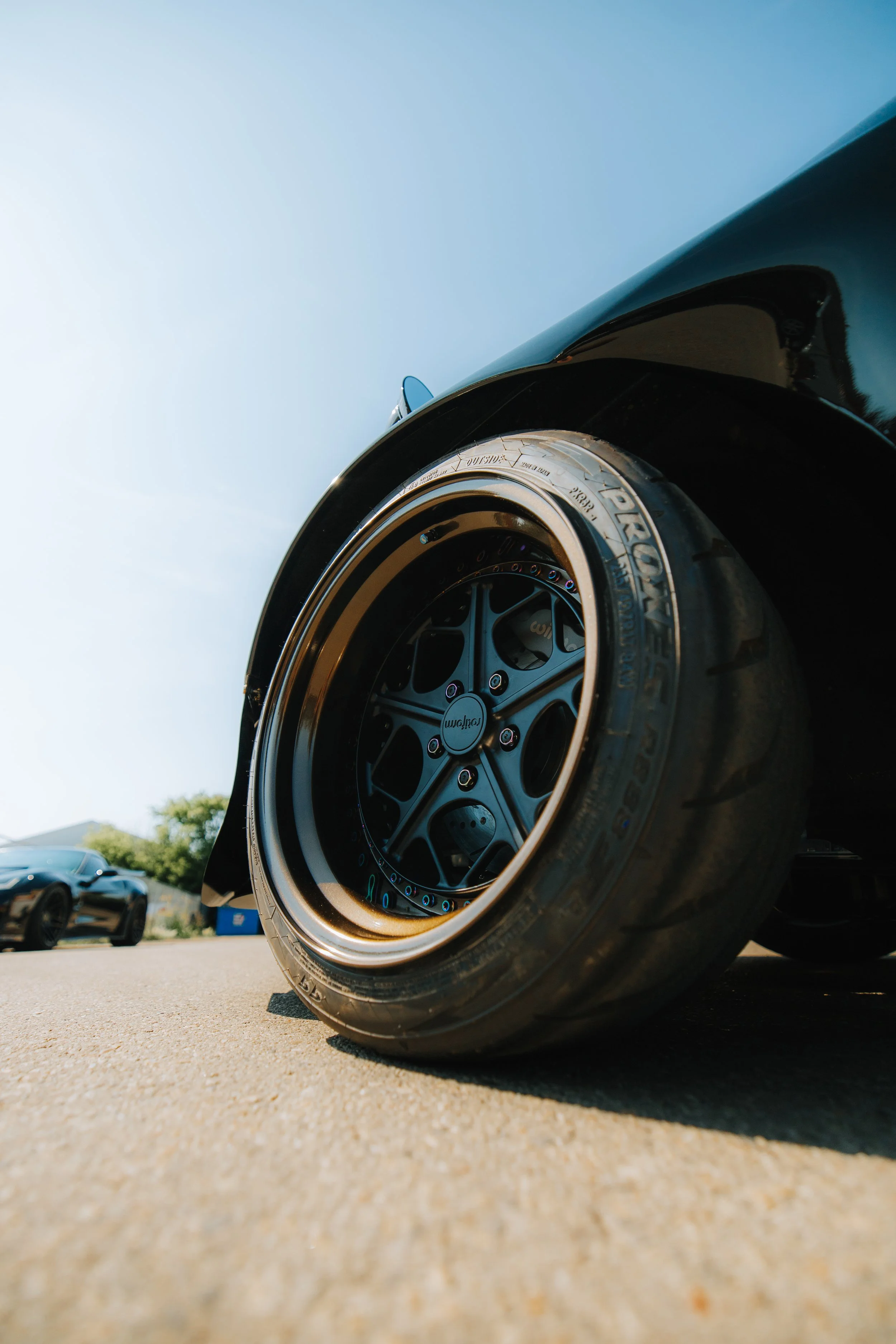 Close-up of a black car's wheel with a gold rim and racing tire, parked on a concrete surface under a clear sky.