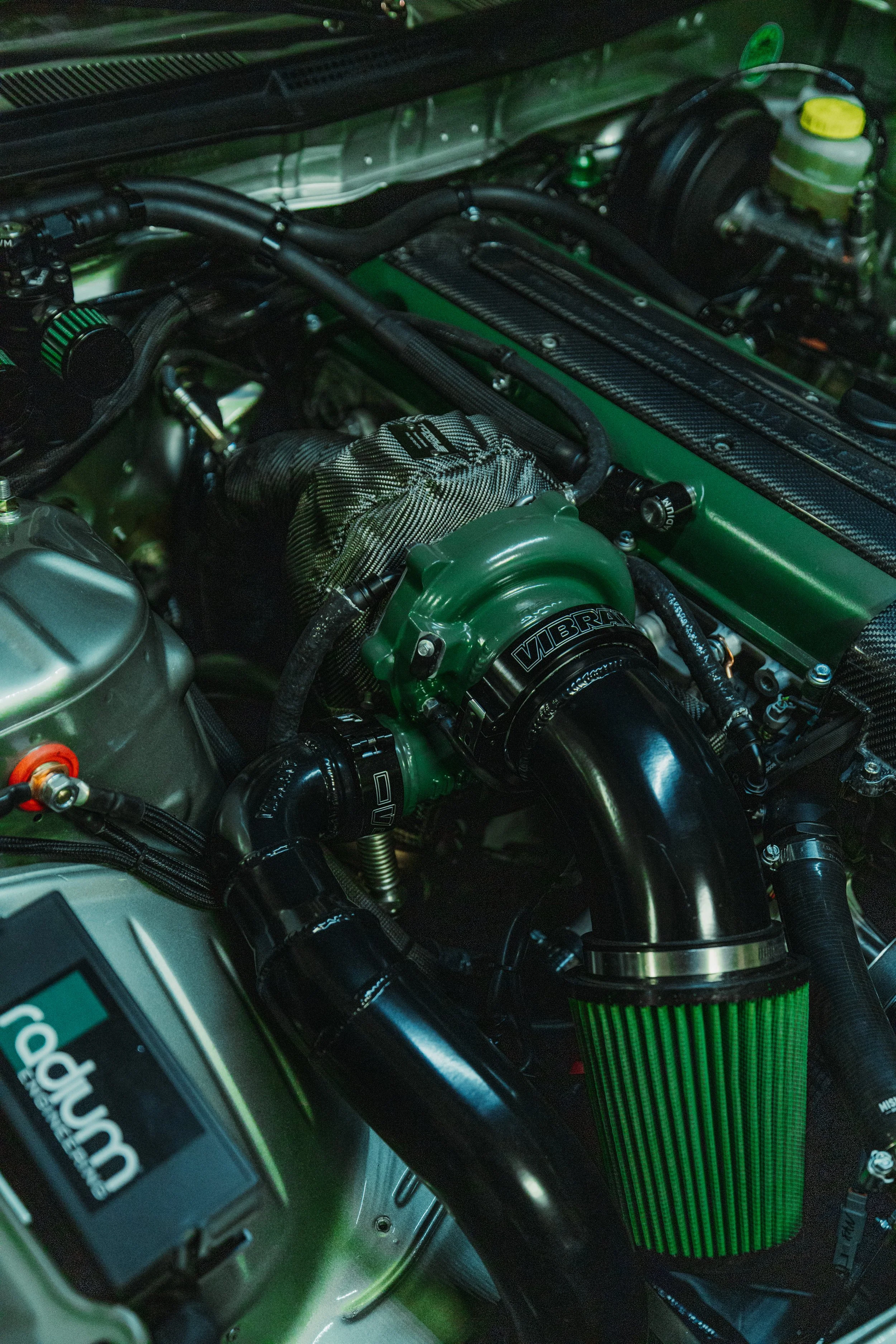 Close-up of a car engine with green and black components, including a green filter, black tubing, and visible branding such as 'VIBRANT'.