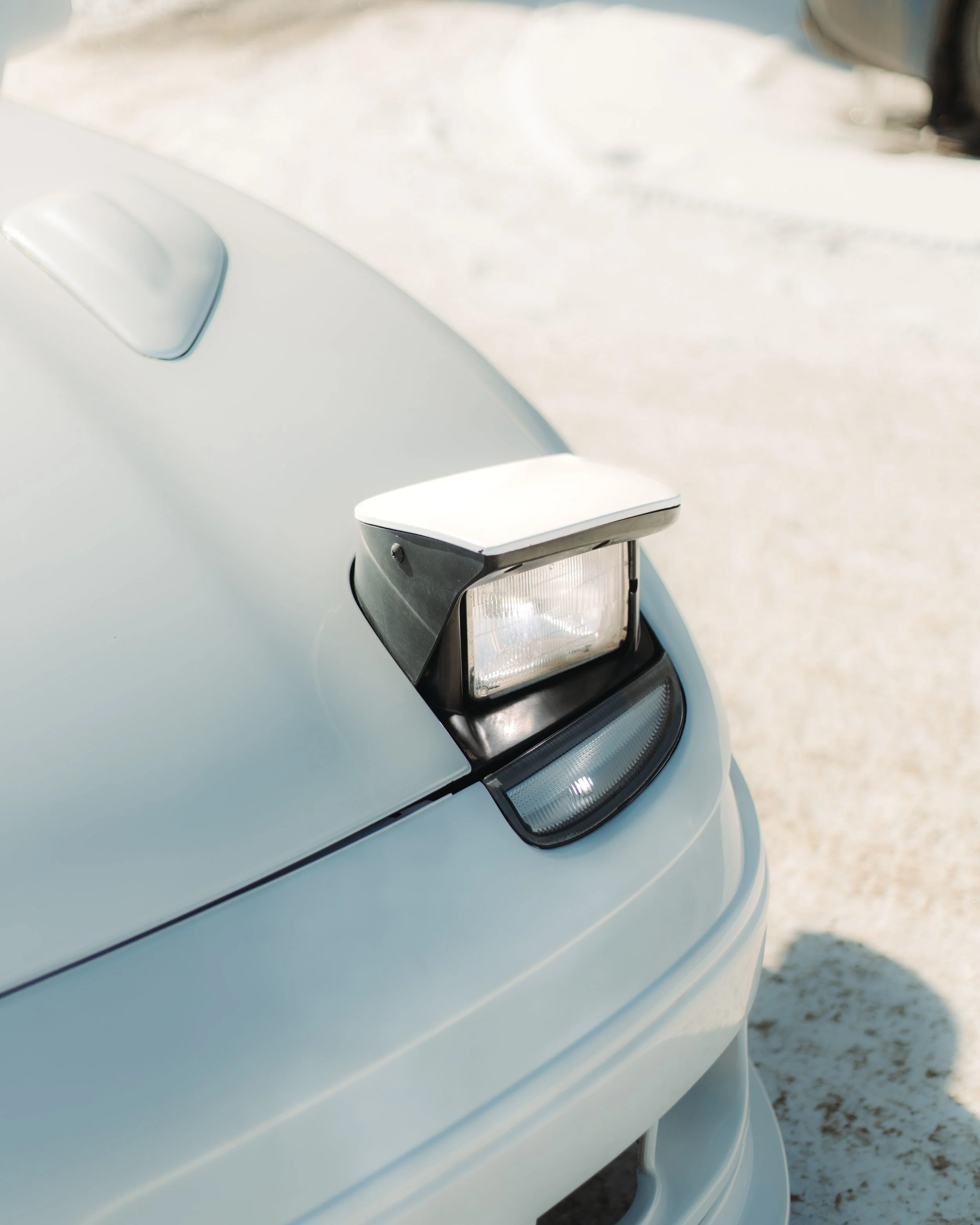 Close-up of a white car's front end with a pop-up headlight raised, on a sunny outdoor surface.