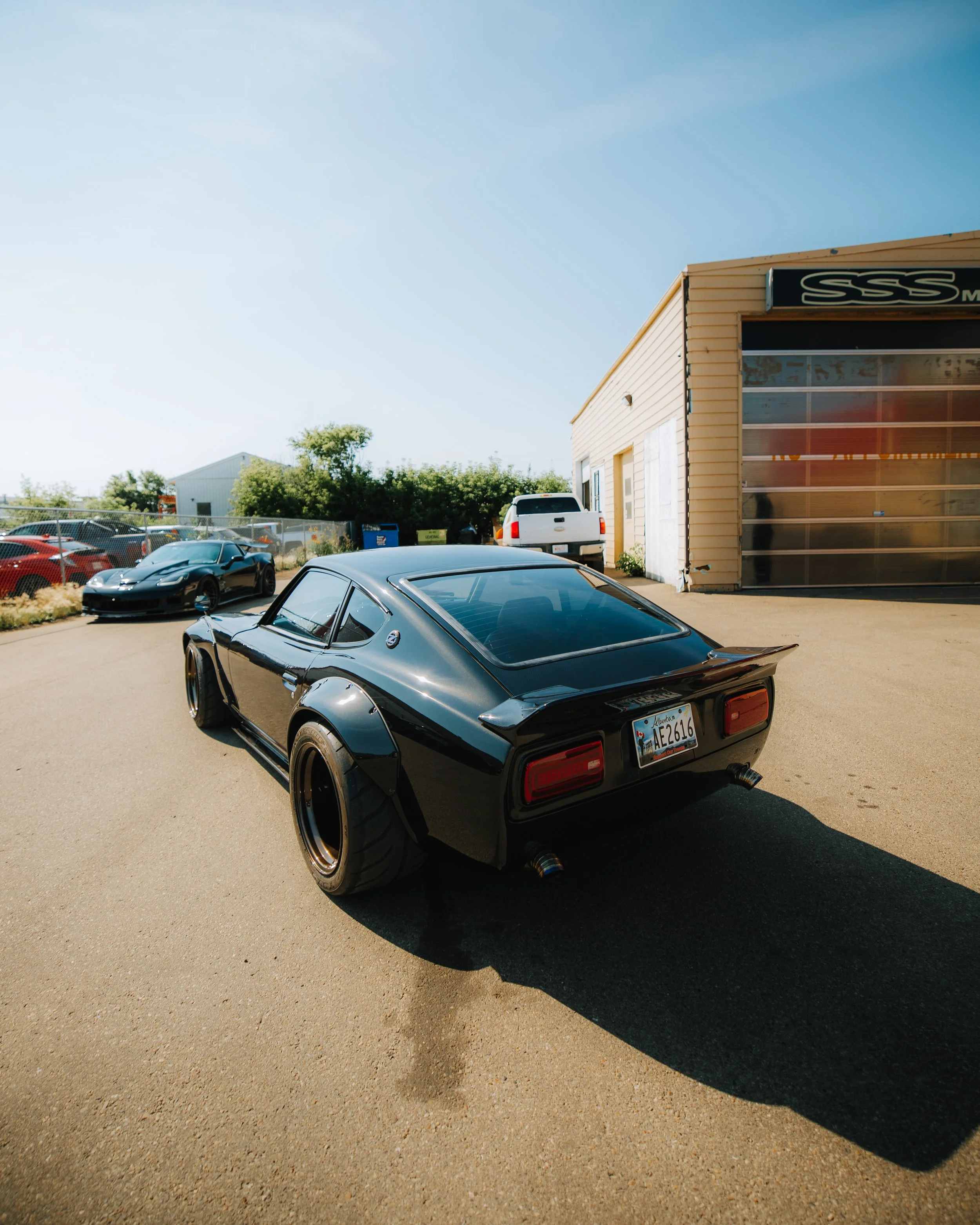 A black, vintage sports car with wide tires parked on a tan asphalt lot near a beige building with a glass garage door, and other cars in the background under a clear blue sky.