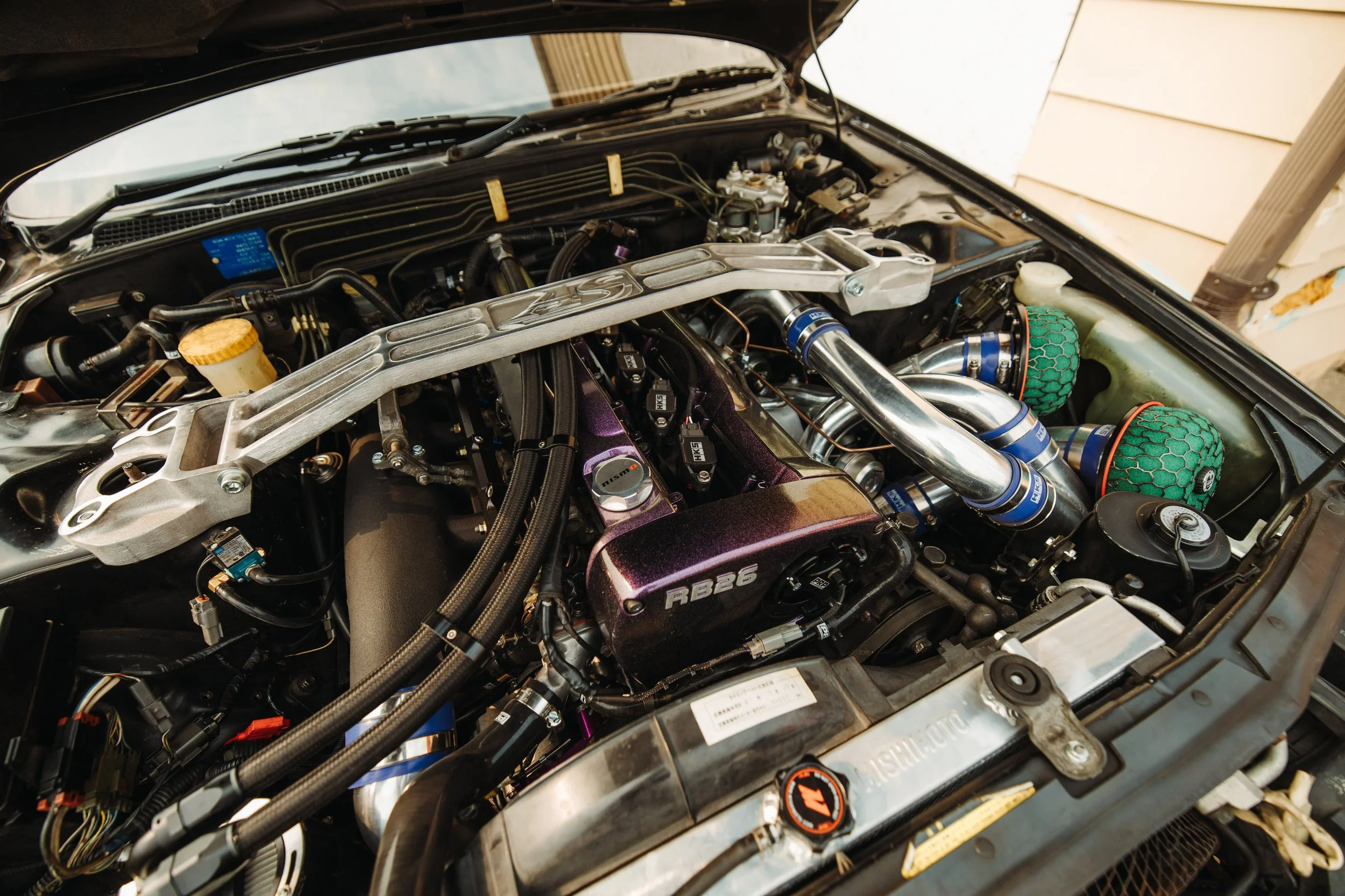 Under the hood of a car, showing a detailed engine with various components, pipes, filters, and a purple cover labeled "RB26".