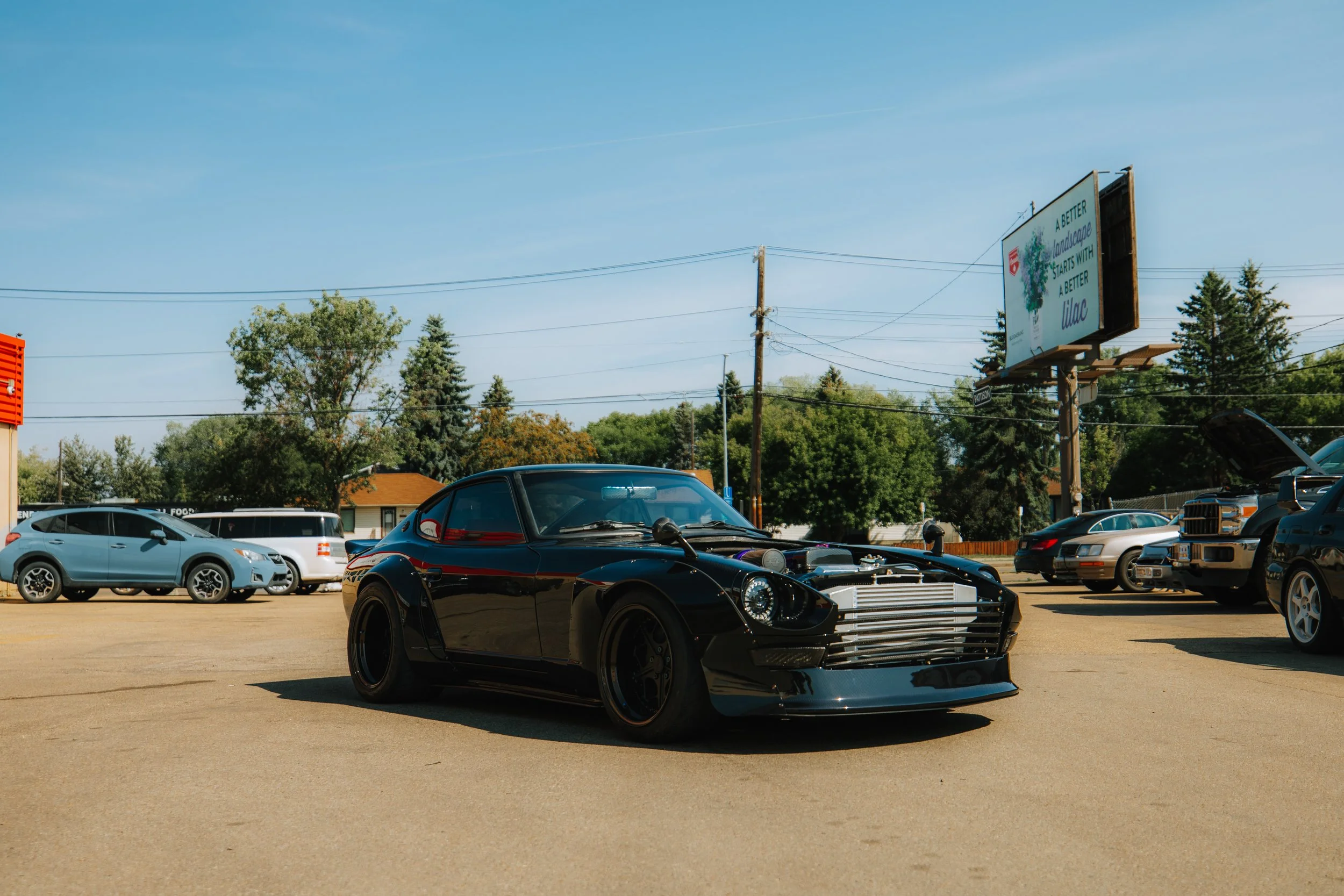 Black vintage sports car with exposed engine and large front grille parked on a lot, surrounded by other cars and trees in the background.