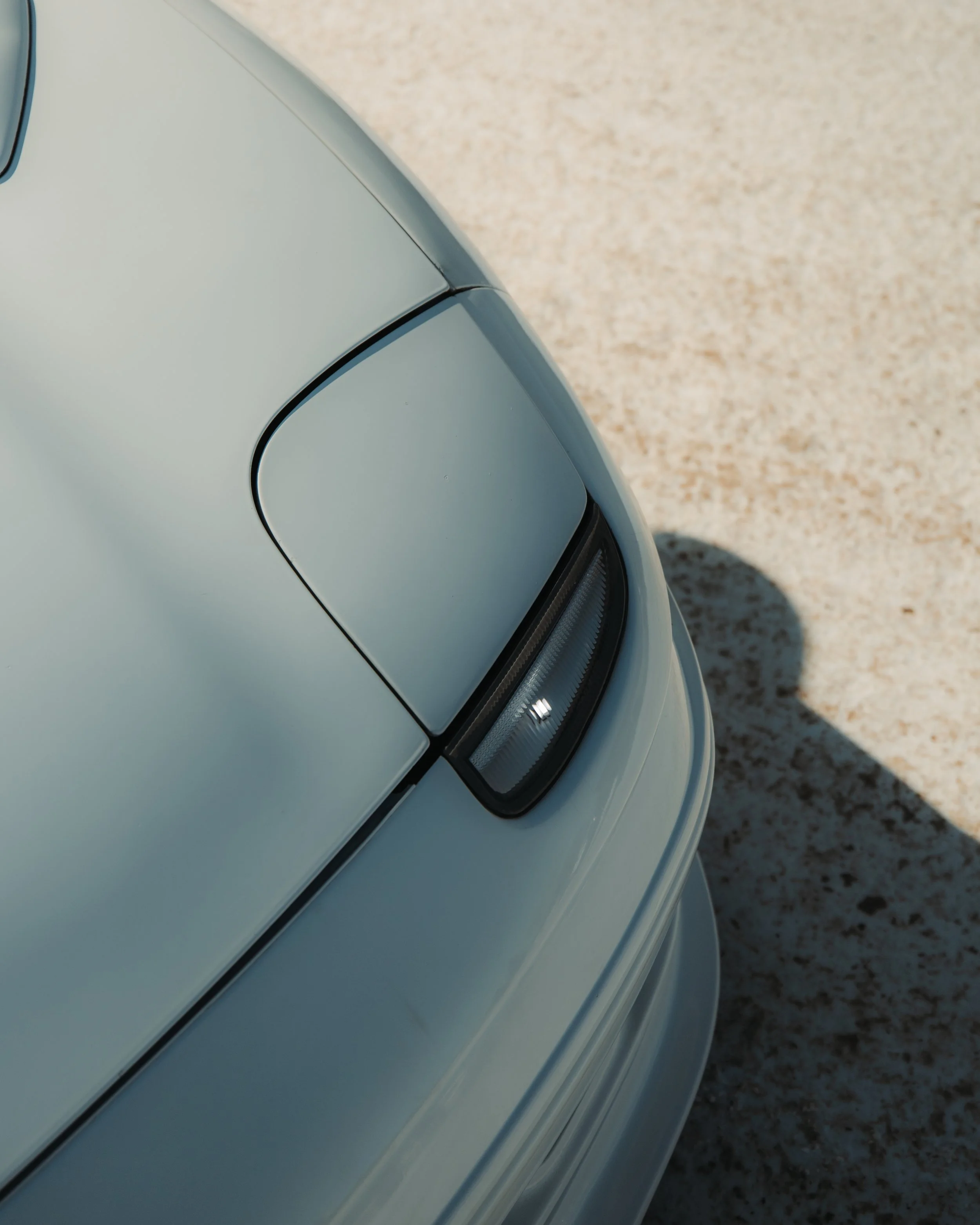 Close-up of the front corner of a light-colored vintage car with a sealed beam headlight and a rounded fender, casting a shadow on a textured light ground surface.