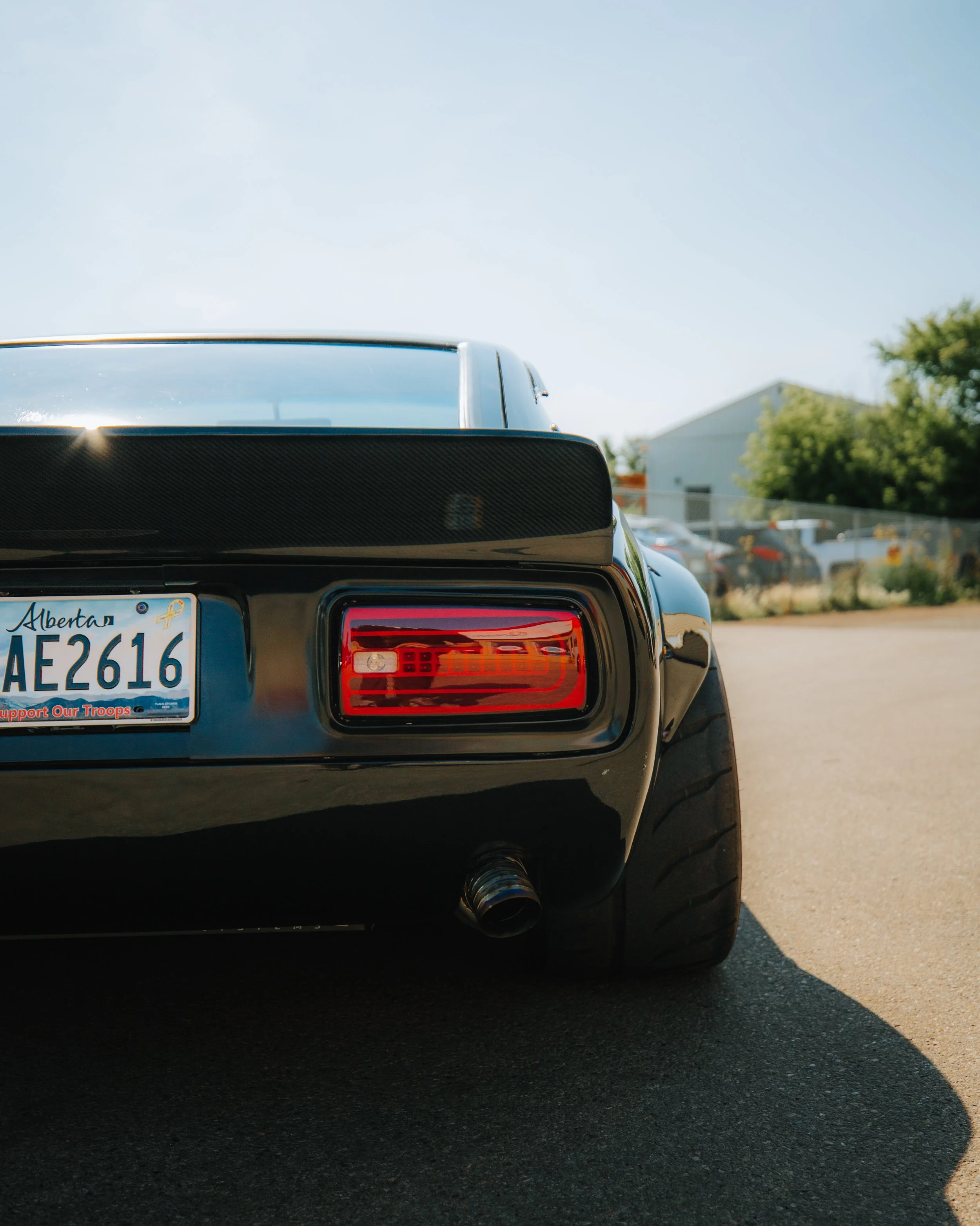 Rear view of a black sports car with a carbon fiber spoiler, showing the right tail light, a custom exhaust pipe, and an Alberta license plate that reads 'AE2616' on a sunny day with a blurred background of a parking lot and trees.