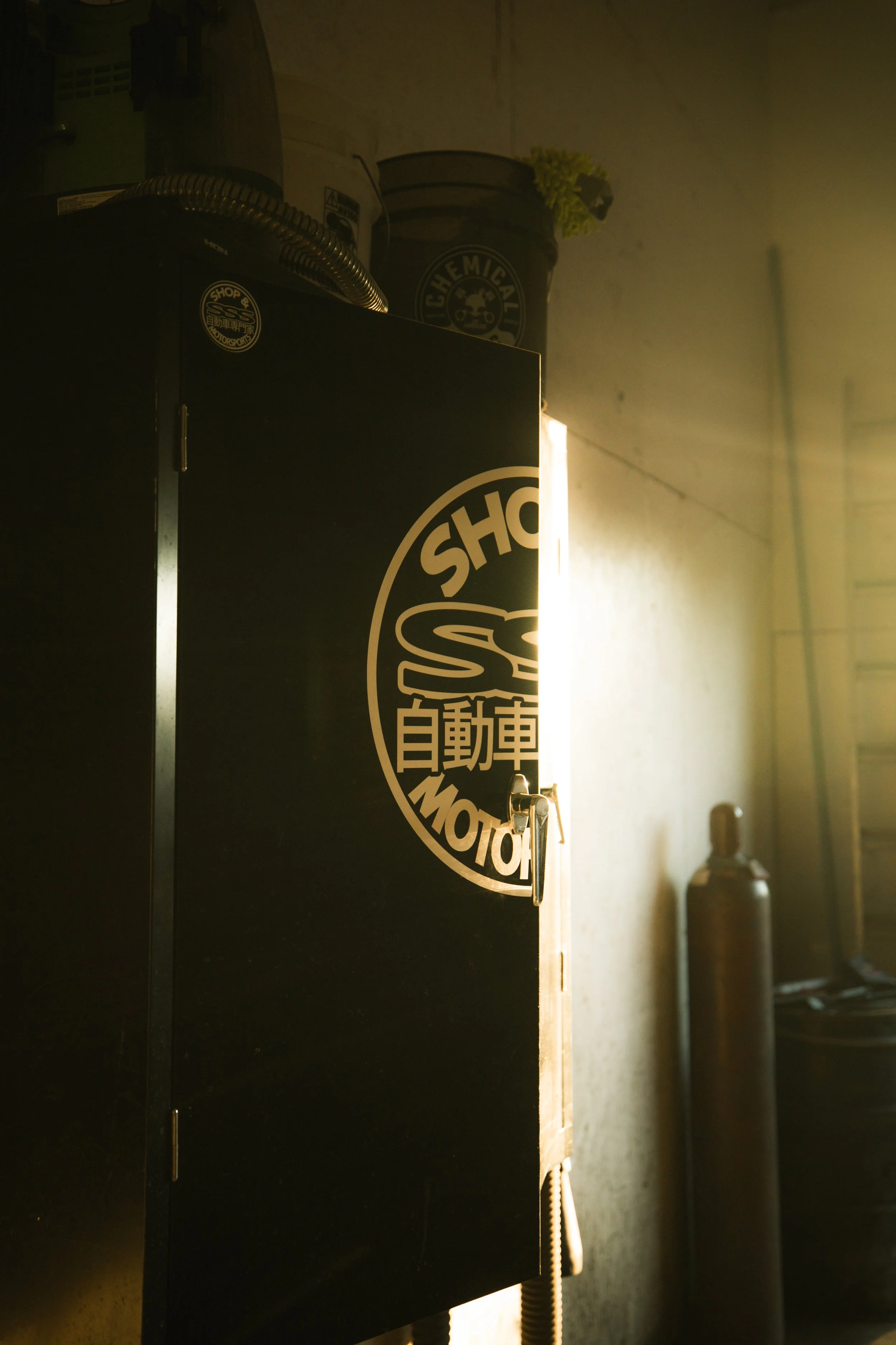 A black metal cabinet with a logo sticker that reads 'Shop SS' and Japanese characters, with sunlight illuminating the side of the cabinet and some items on top, including a bucket and a propane tank in a garage or workshop setting.