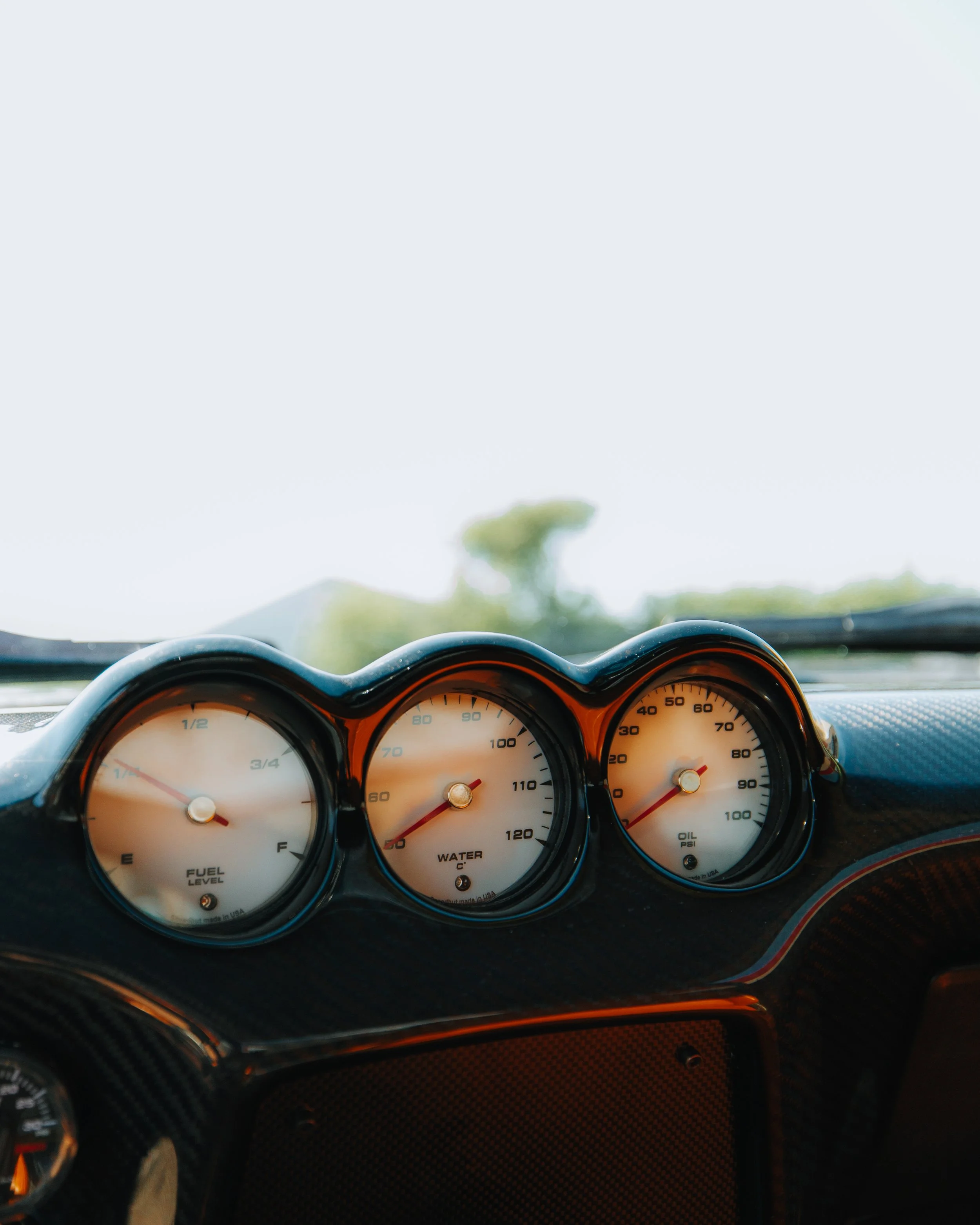 Close-up view of a car dashboard with three round gauges showing fuel level, water temperature, and oil pressure, with a blurred outdoor background.