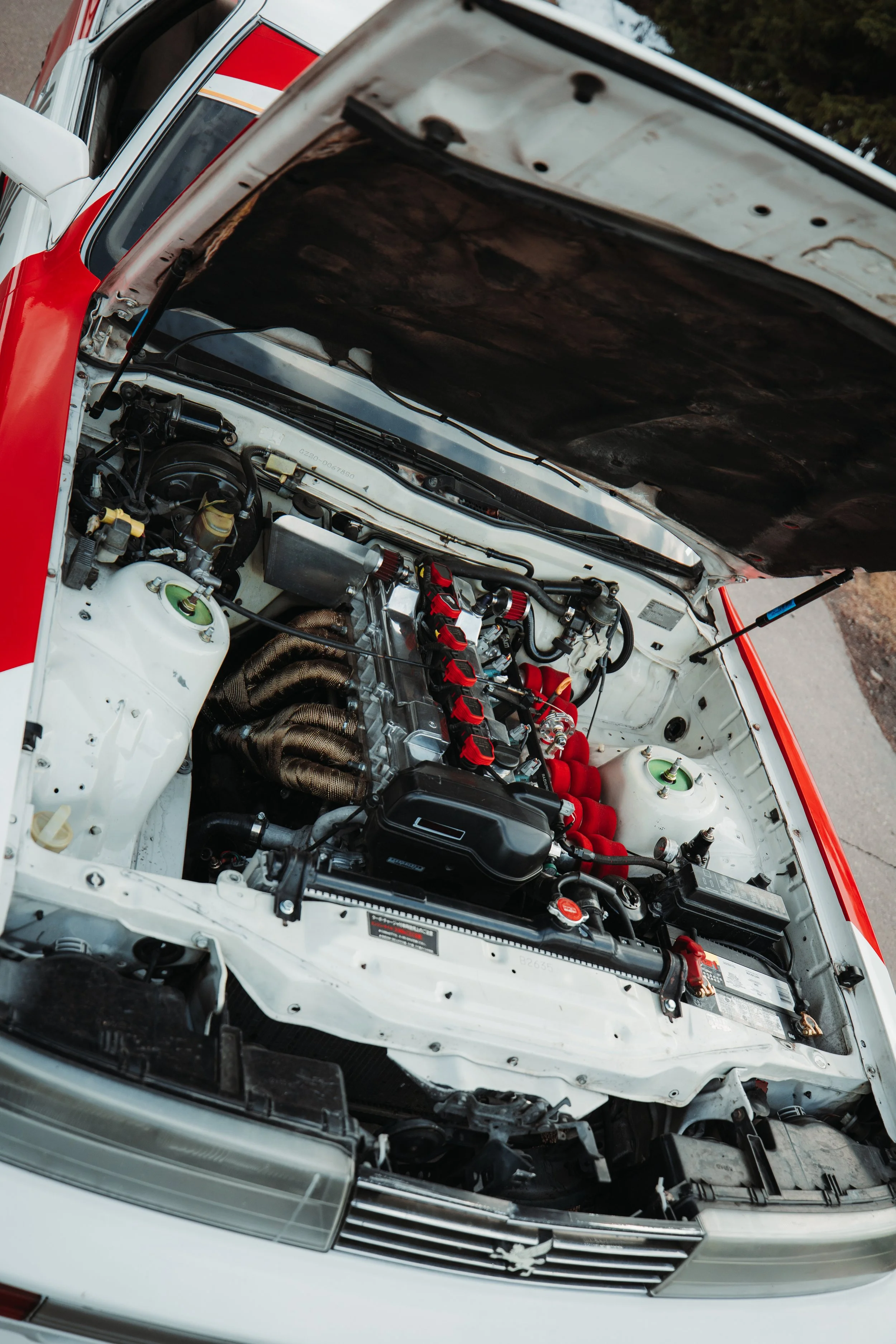 Close-up view of a car engine with the hood open, showing various engine components, hoses, and wiring inside a white engine bay with some red accents.