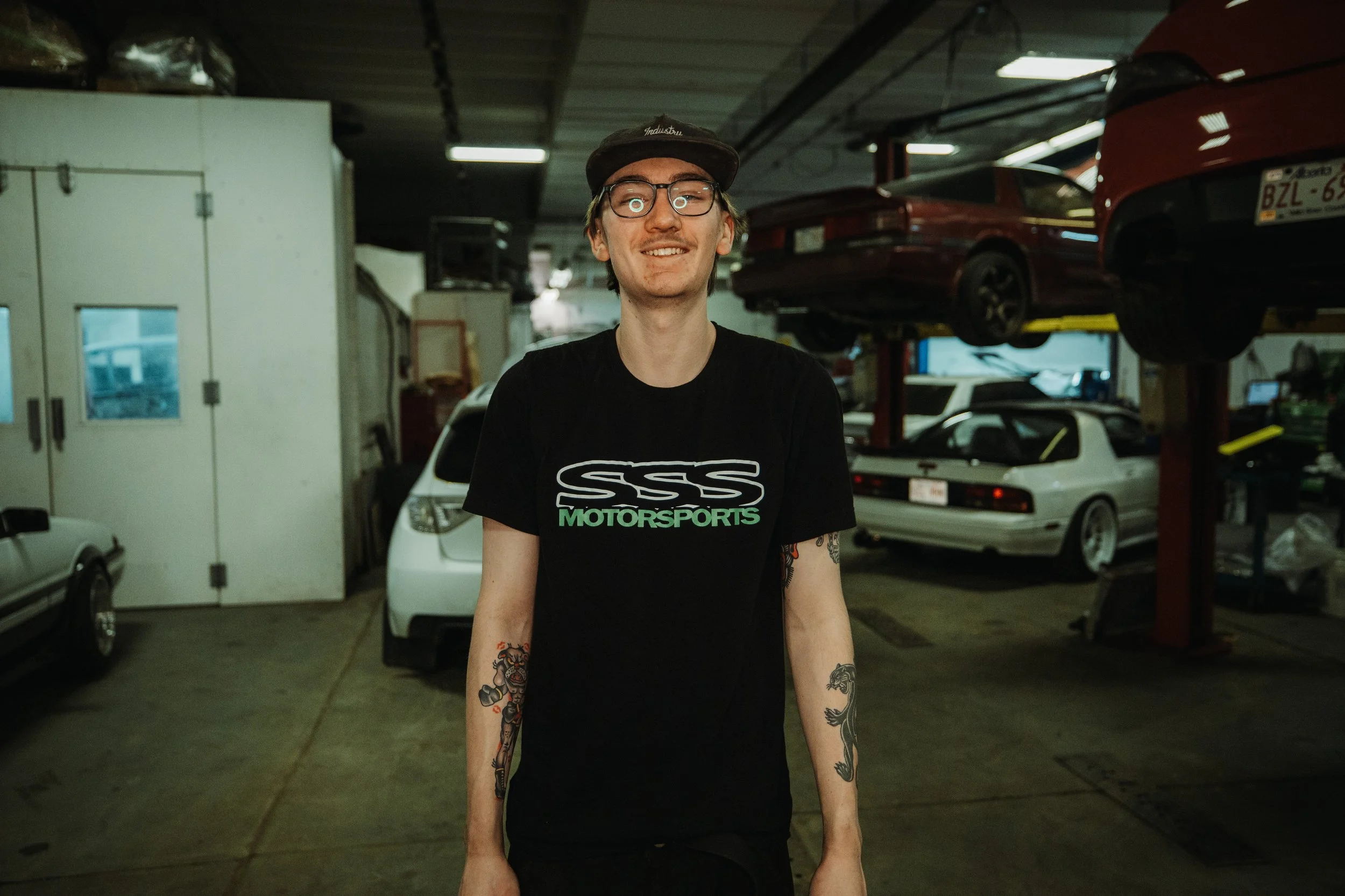 A young man with glasses, a cap, and tattoos on both arms standing inside a garage surrounded by parked cars in various stages of maintenance or repair.