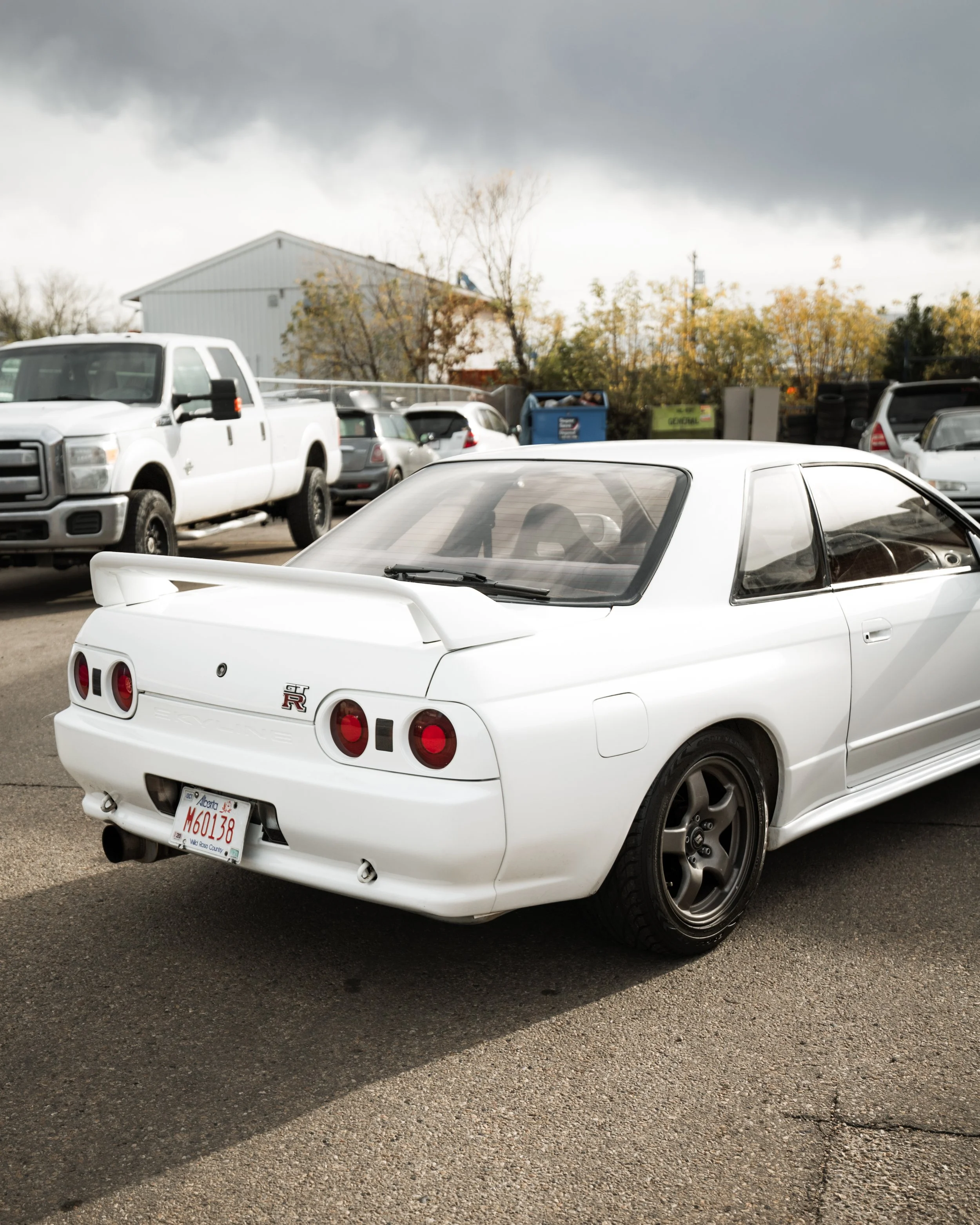 A white vintage Nissan Skyline GT-R R32 sports car parked outdoors with other vehicles and a cloudy sky in the background.