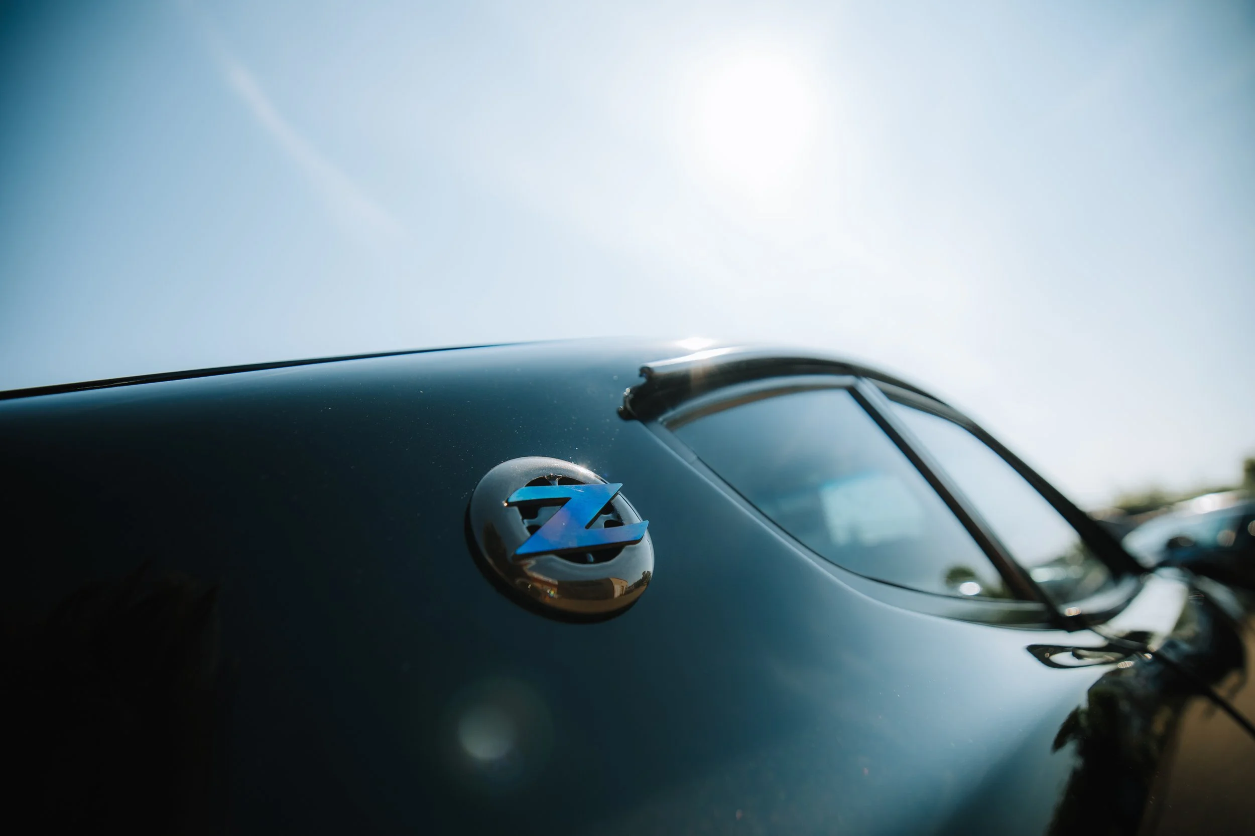 Close-up of a vintage black car with a chrome badge featuring a blue 'Z' emblem on the hood, under a bright sky with the sun.