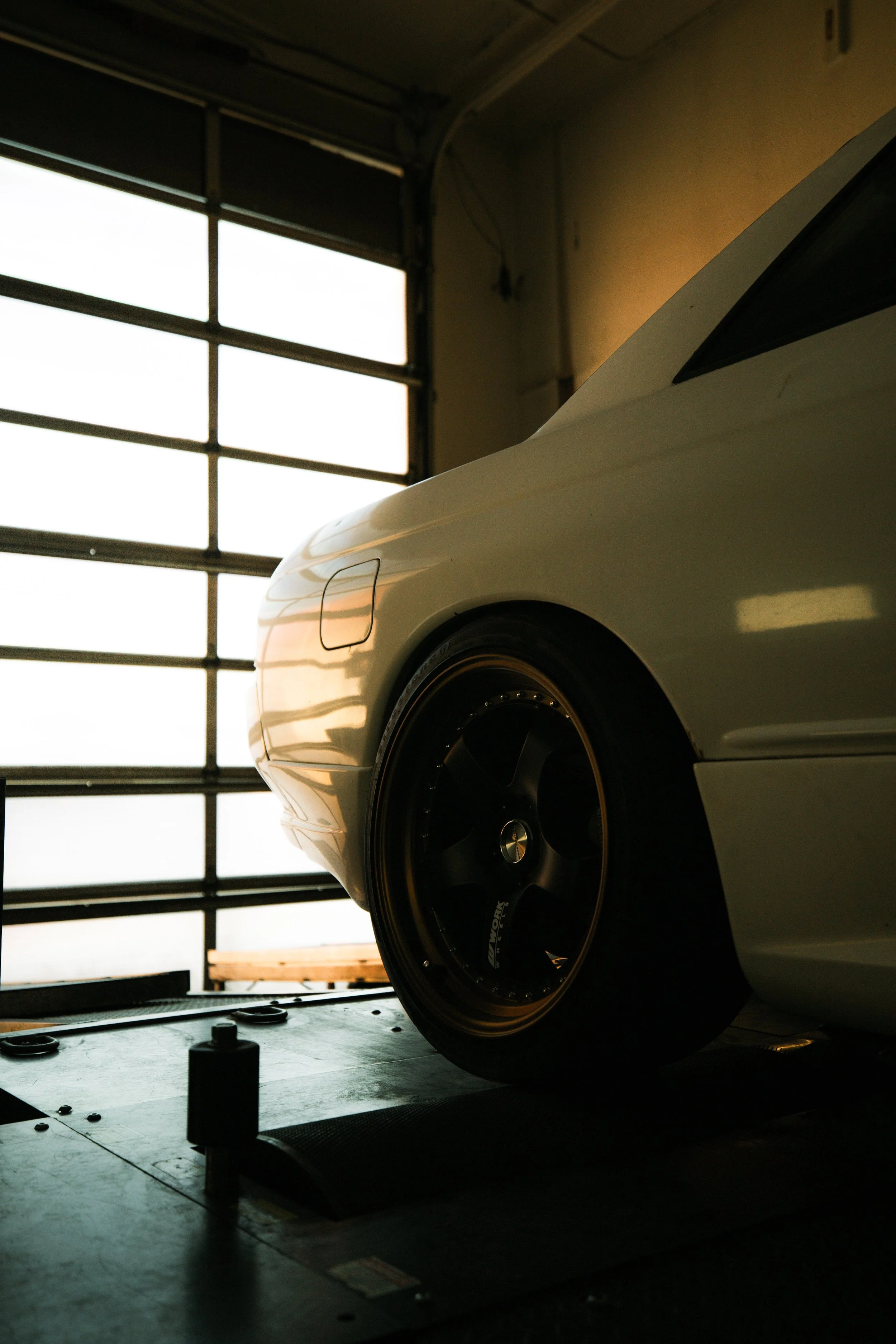 Part of a white sports car in a garage, with focus on the front wheel and fender, and a large garage door in the background.