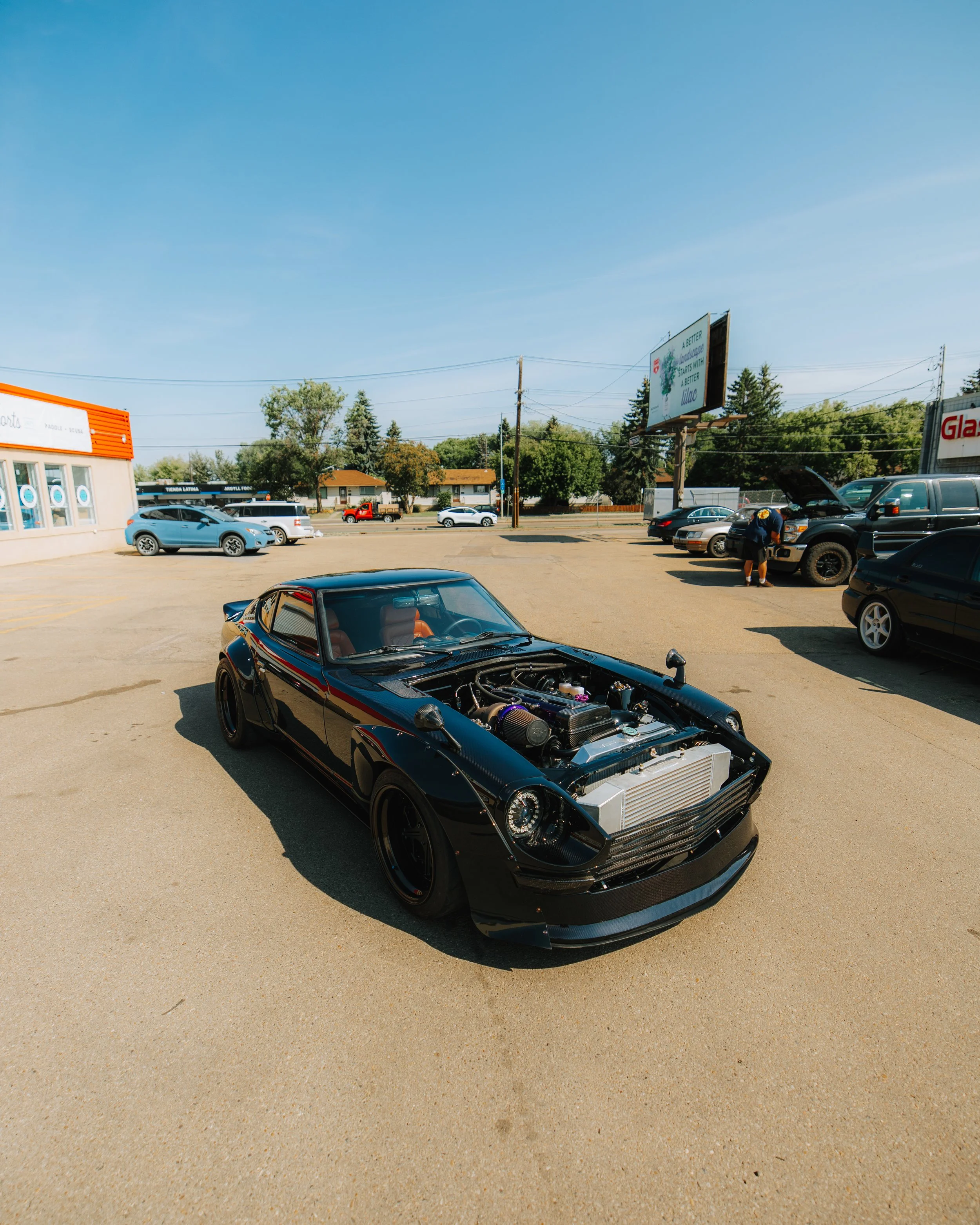 A black sports car with its hood open parked in a lot, with other vehicles and a billboard in the background under a blue sky.