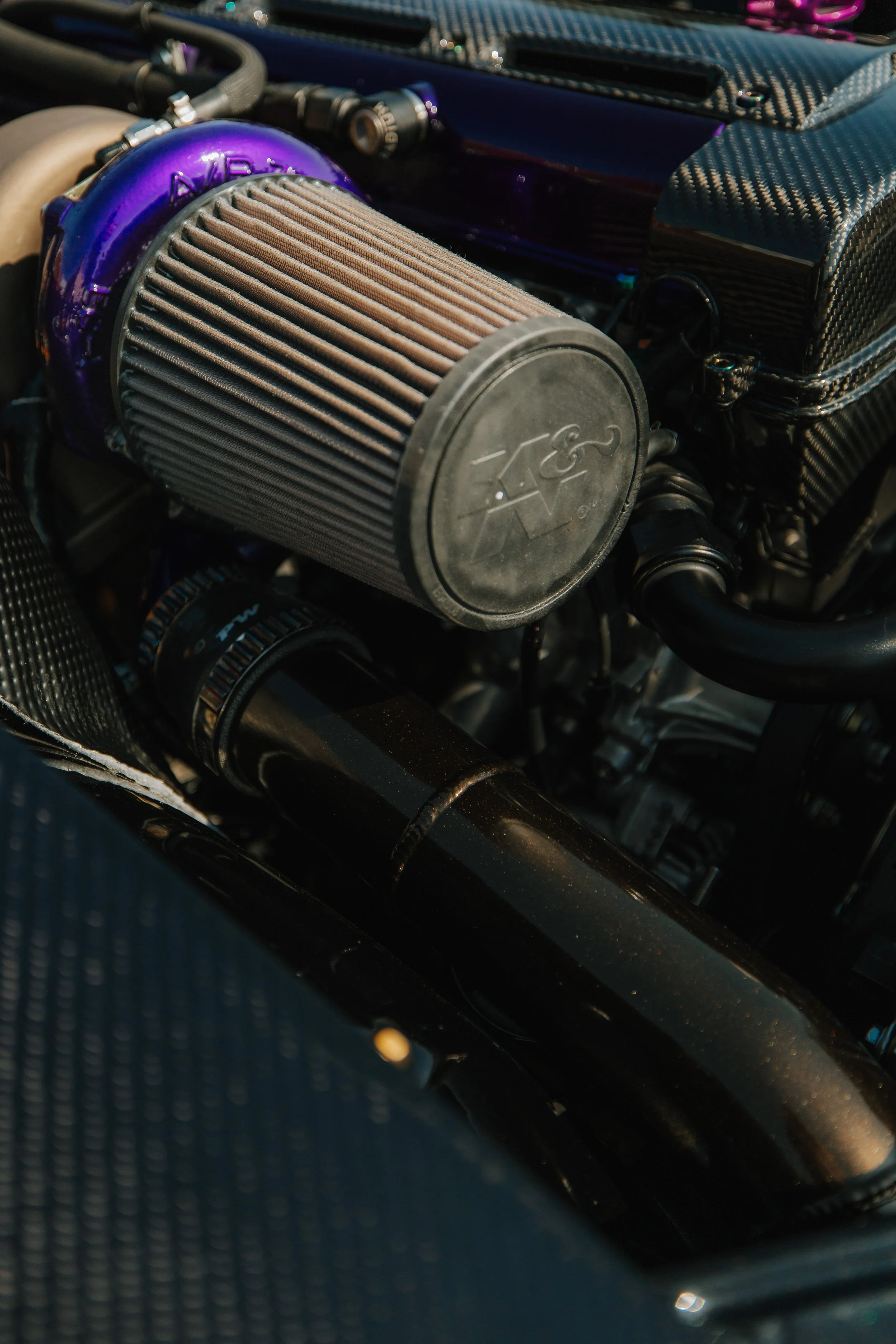 Close-up of a high-performance car engine with a large cone-shaped air filter labeled with an automotive logo, black tubing, and carbon fiber components.