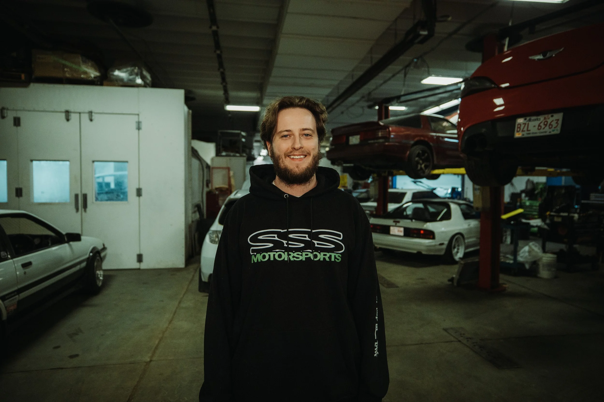 A smiling young man with a beard and brown hair standing in a garage filled with classic cars, some on lifts and others parked on the floor.