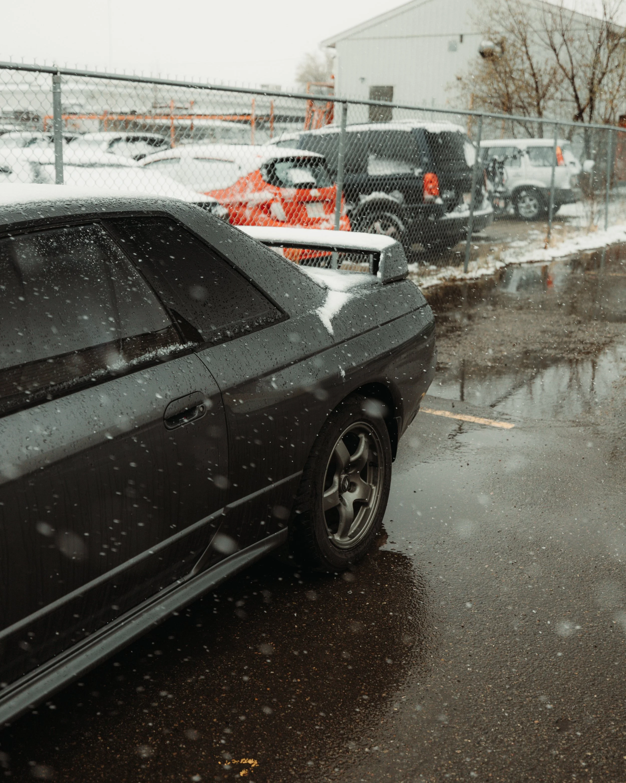 A black sports car with rain droplets on it, parked on a wet asphalt surface near a snow-covered lot with other vehicles and a chain-link fence in the background.