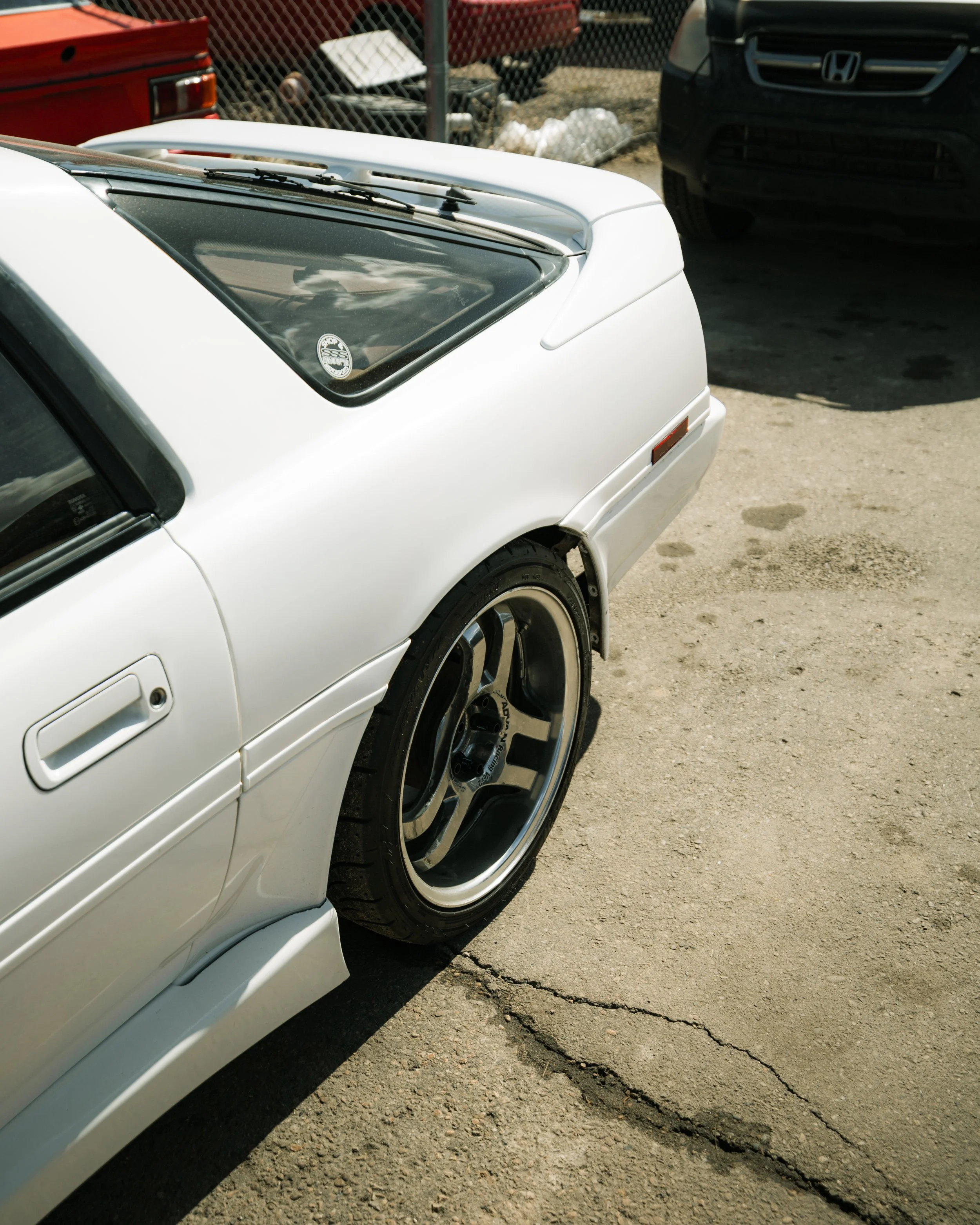 Close-up of the rear side of a white vintage sports car with a black wheel and tire, parked on a cracked concrete surface with other cars and a chain-link fence in the background.