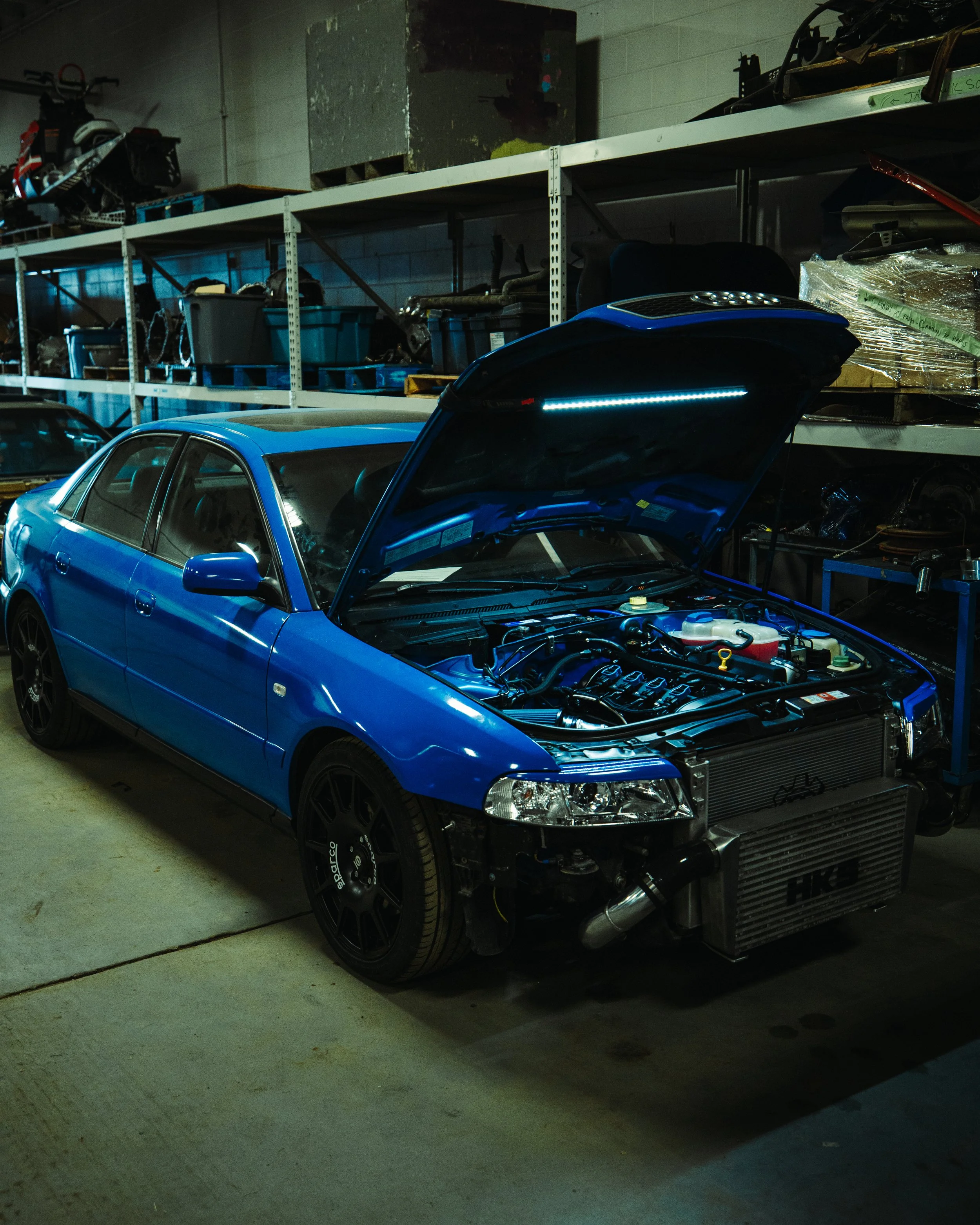 Blue car with its hood open, showing the engine inside, parked inside a garage. The garage has shelves with various car parts and tools.