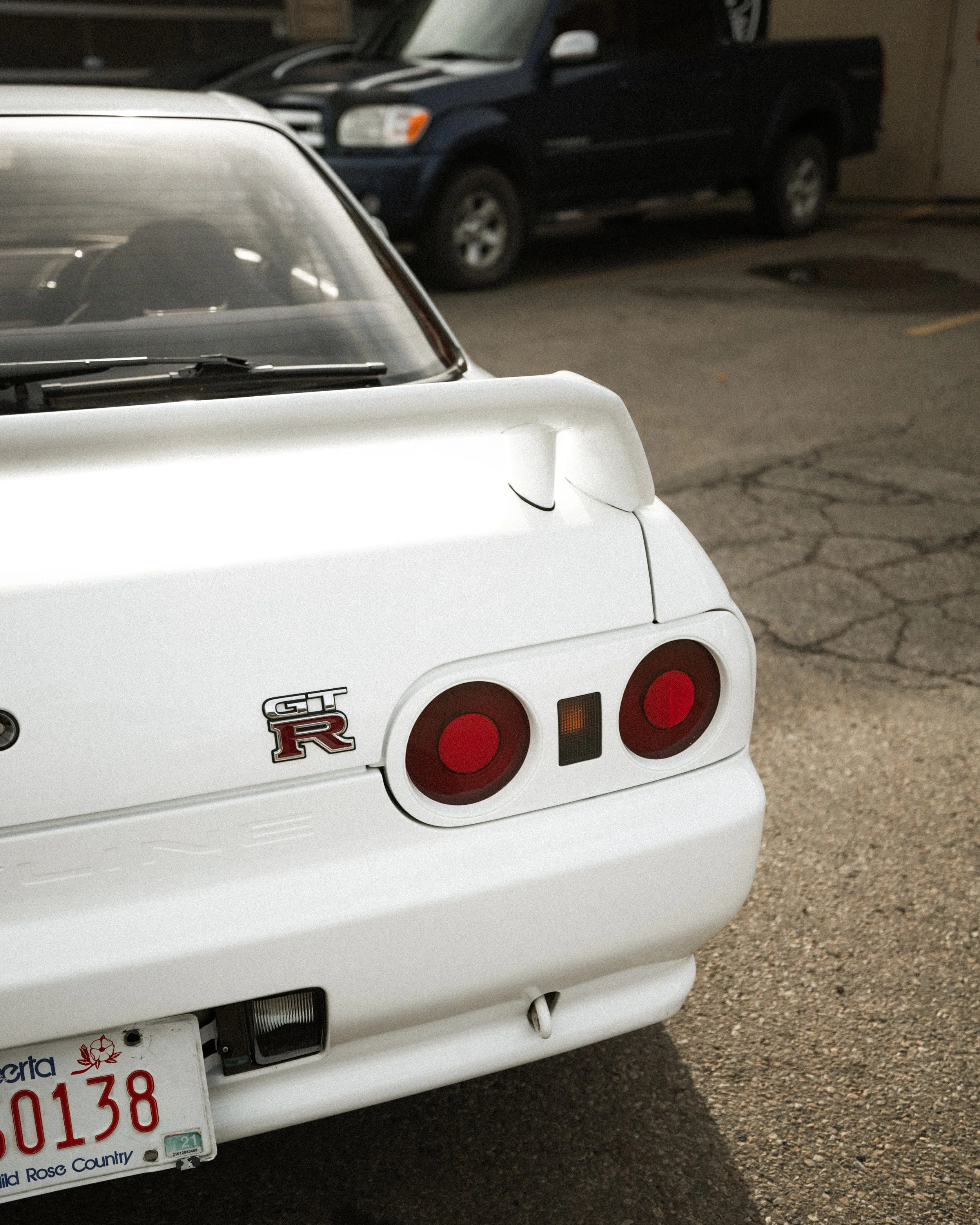 Close-up of the rear of a white Nissan GT-R sports car, showing the GT-R badge, round tail lights, and a partial view of a license plate, parked in a lot with other vehicles.
