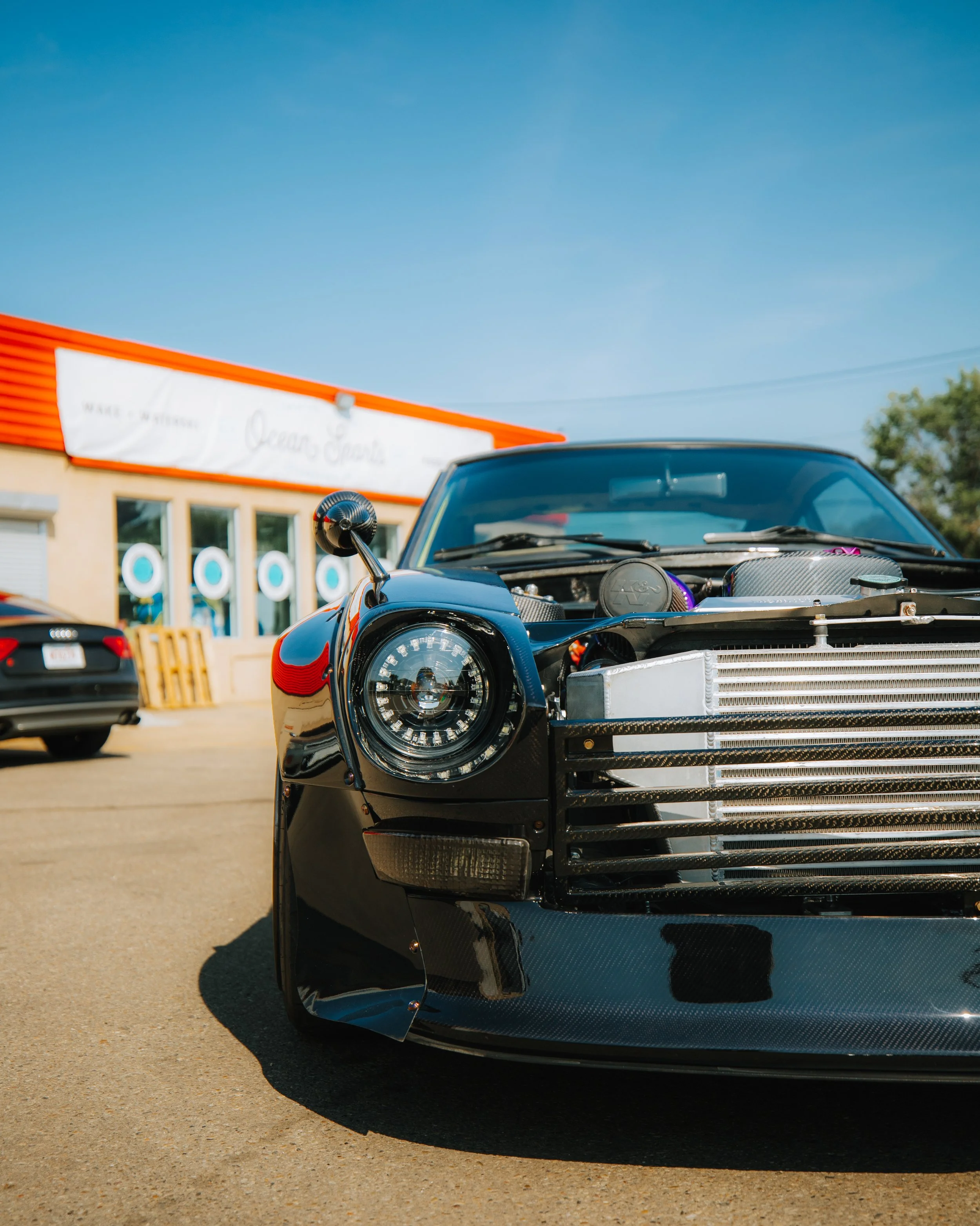 Close-up of a black sports car with an open hood, exposing the engine, parked outside a building with a sign that reads 'Ocean Dive' under a clear blue sky.