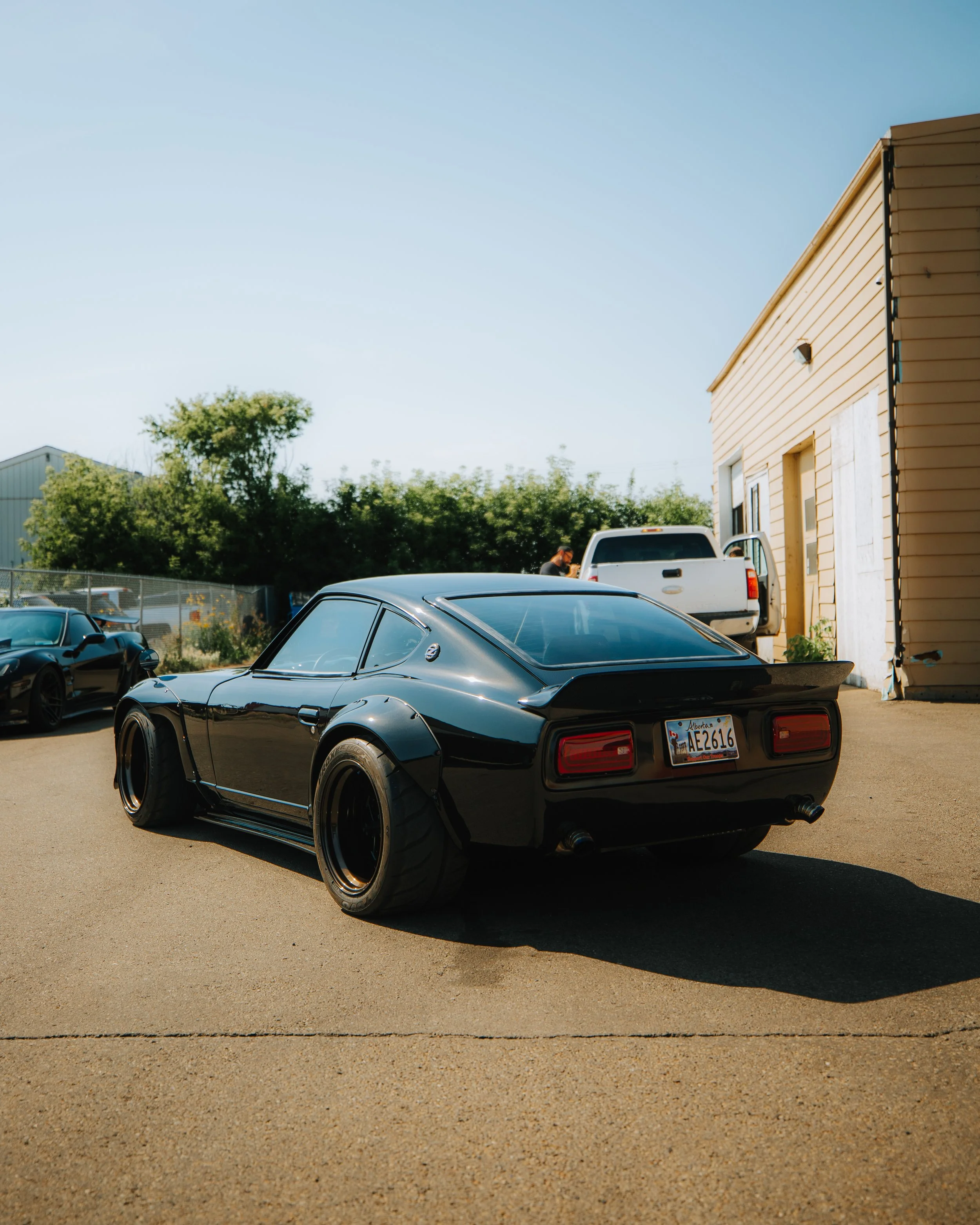A black sports car with wide tires and a rear spoiler parked on a concrete surface near a beige building with a white door. Other vehicles and trees are visible in the background under a clear sky.