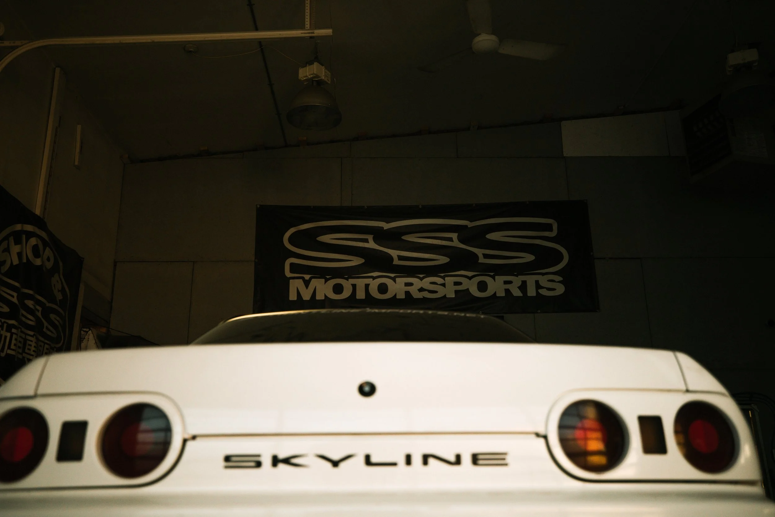 Rear view of a white Nissan Skyline car in a garage with a "SSS MOTORSPORTS" banner hanging on the wall behind it.