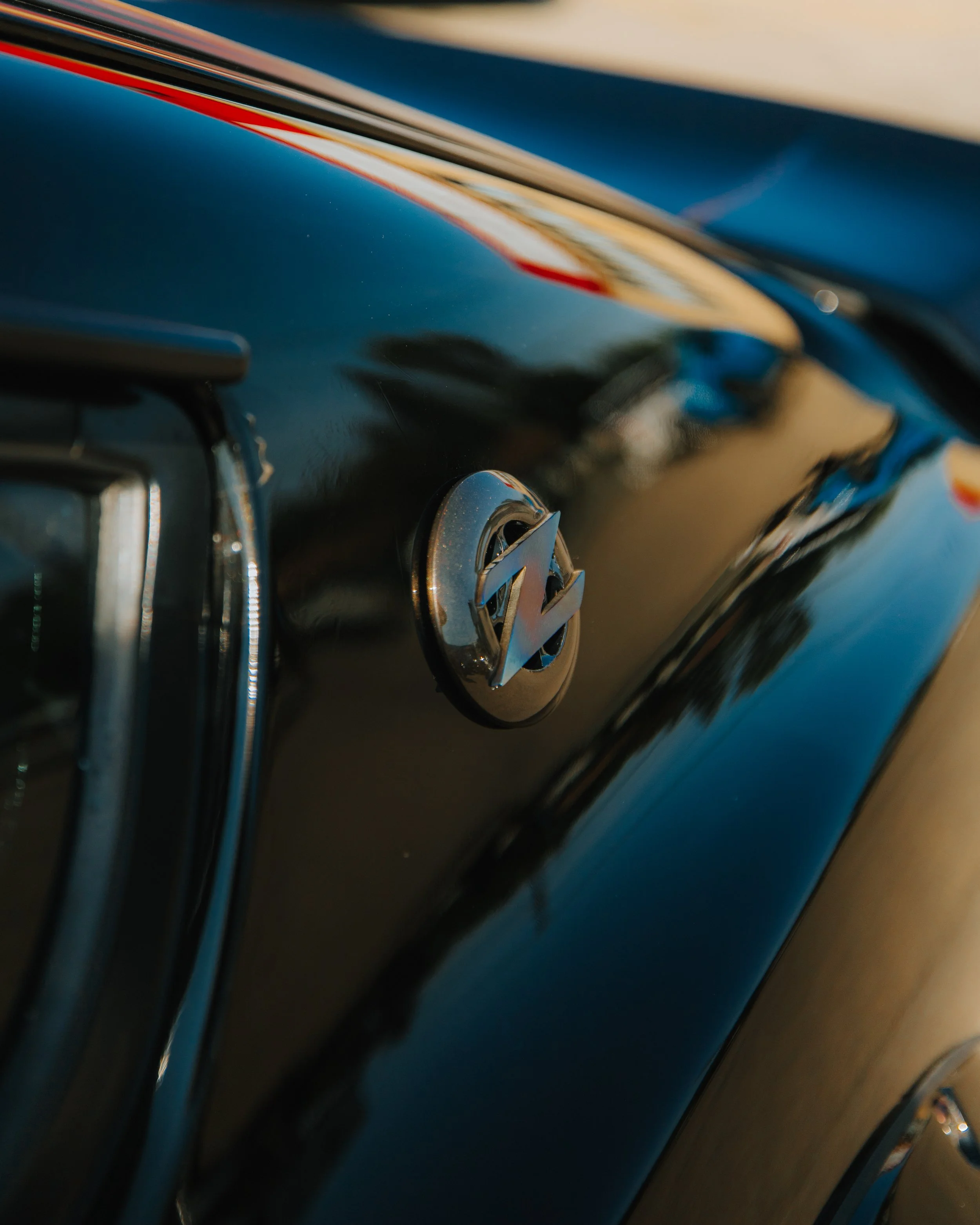 Close-up of a vintage car's hood ornament featuring a stylized 'V' logo, with a black glossy surface reflecting the surroundings.