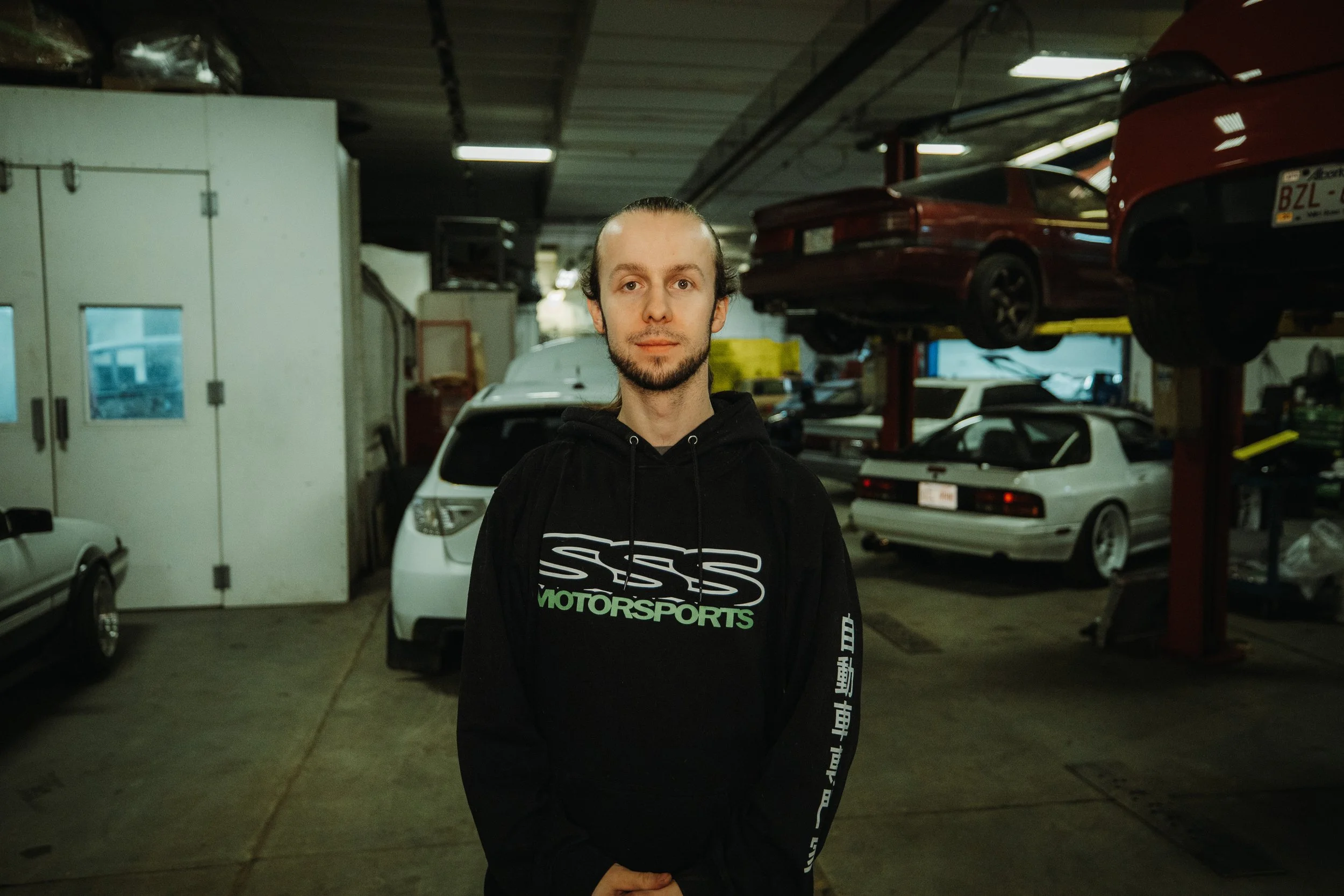A young man with long hair and a beard standing in a garage or auto repair shop, wearing a black hoodie with white and green graphics and text, surrounded by cars and car lifts.