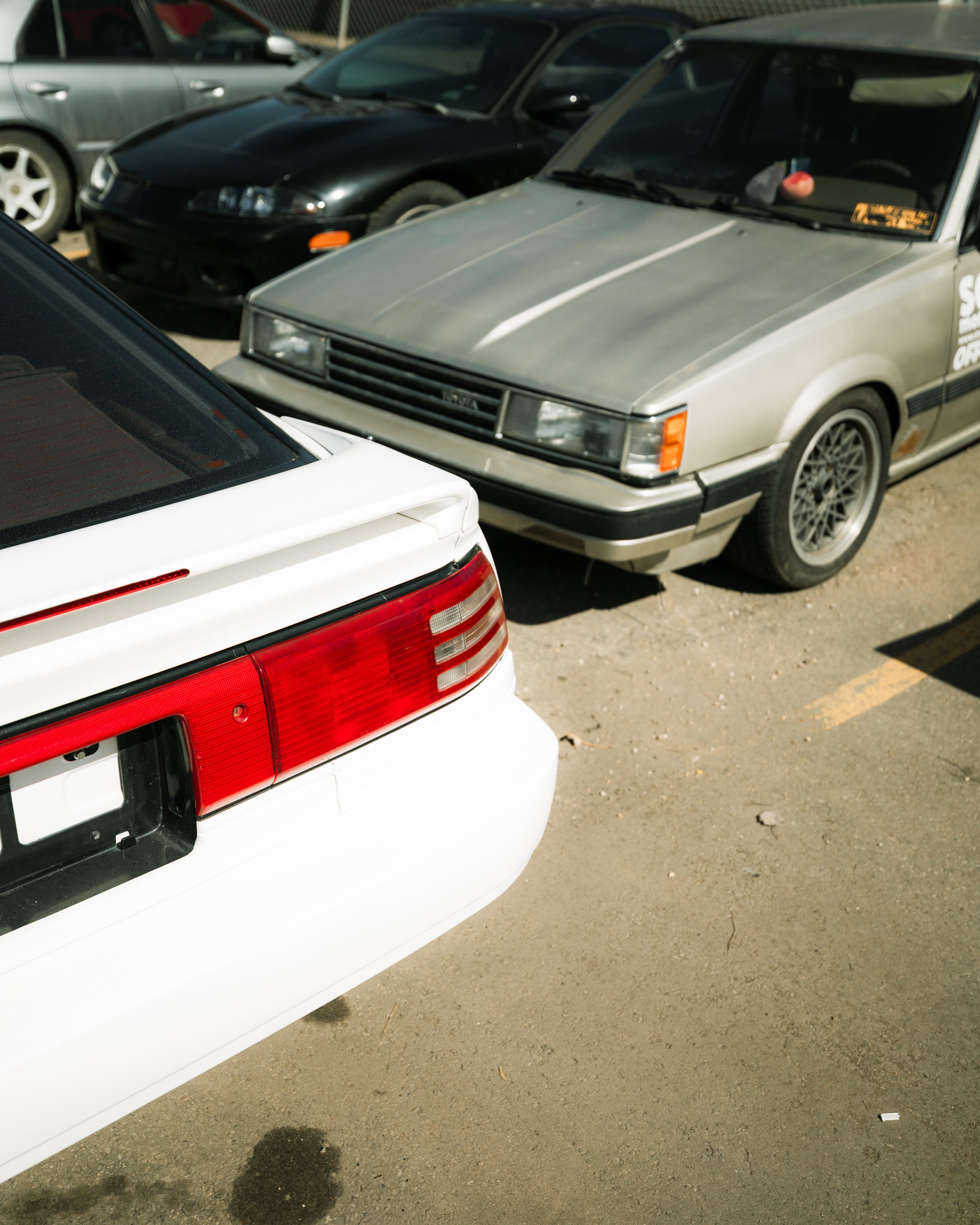 Several cars parked in a lot, including a close-up of a white car's rear with red tail lights, a black car in the middle, and a gray car with a slightly dusty appearance.