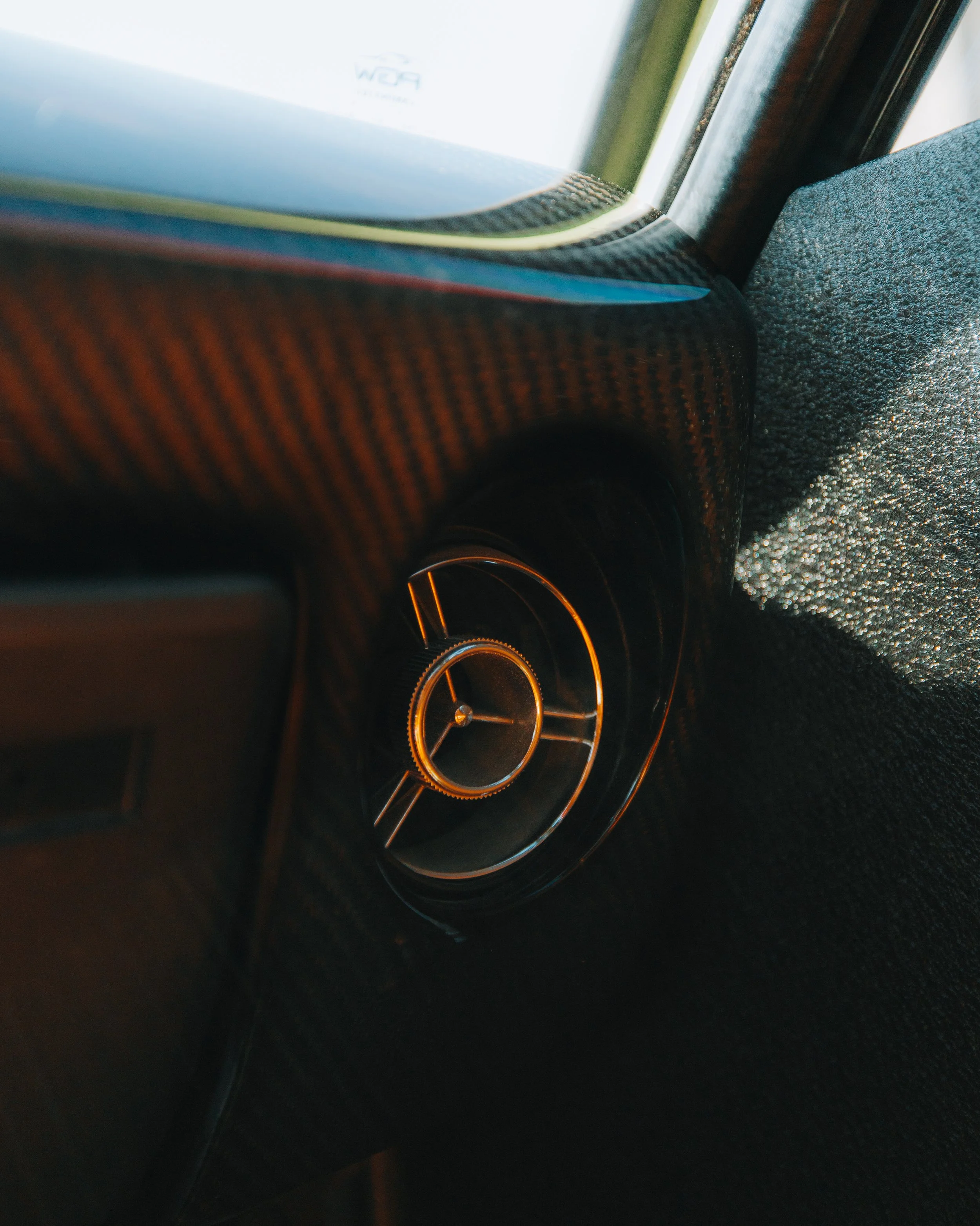 Close-up of a car's circular air vent with a metallic rim, integrated into a carbon fiber dashboard.