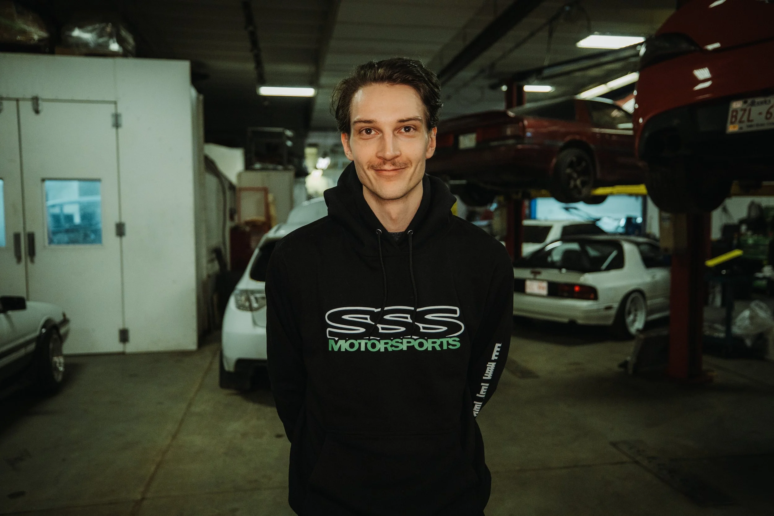 A young man standing in a garage with cars on lifts in the background, wearing a black hoodie with 'SSS MOTORSPORTS' logo.